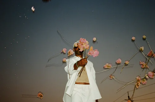 A portrait photograph shot on 35mm film shows a person wearing a white blazer and crop top against a twilight sky, holding dried flowers and branches with faded pink and brown blooms.