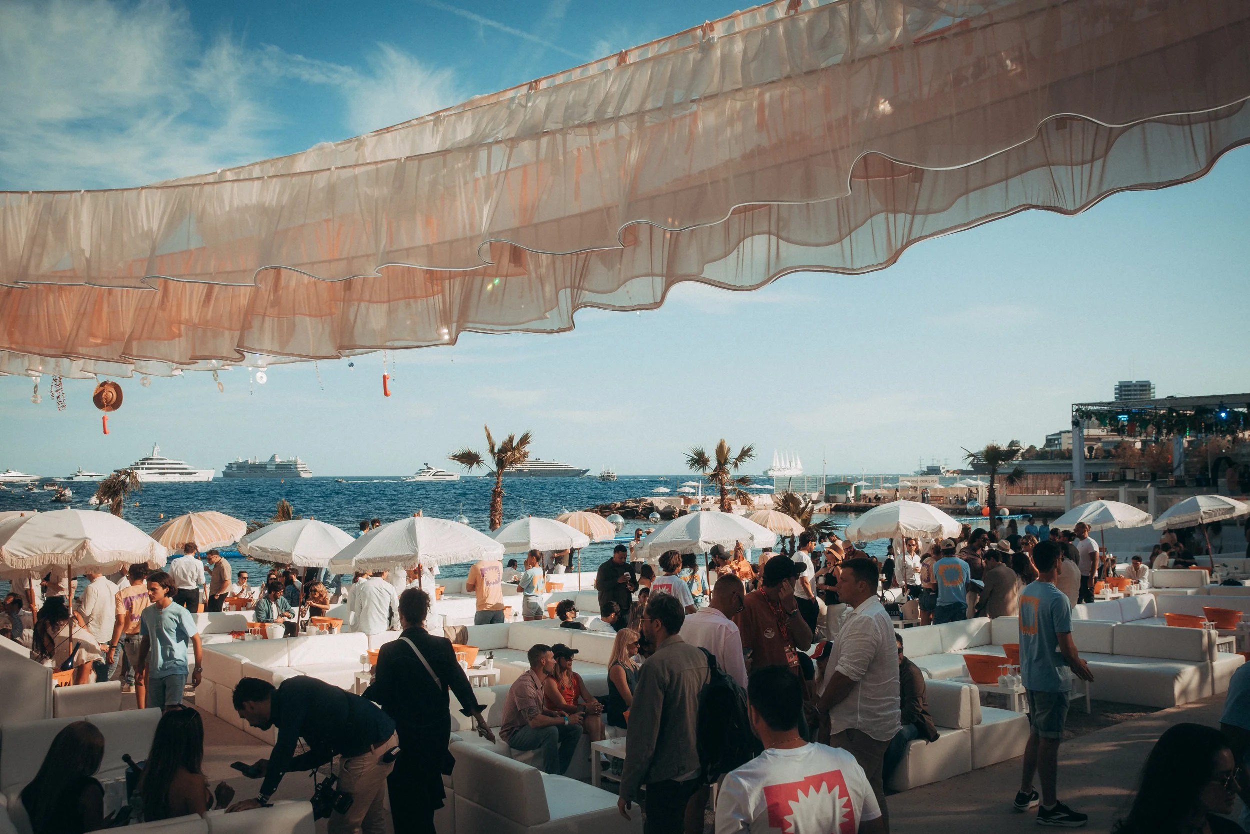 People gathering at a beachside outdoor lounge with white sofas, umbrellas, and palm trees, overlooking the ocean with boats and yachts in the distance under a partly cloudy sky.