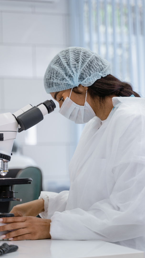 A person wearing a lab suit, a face mask, and hair cap is looking into a microscope.