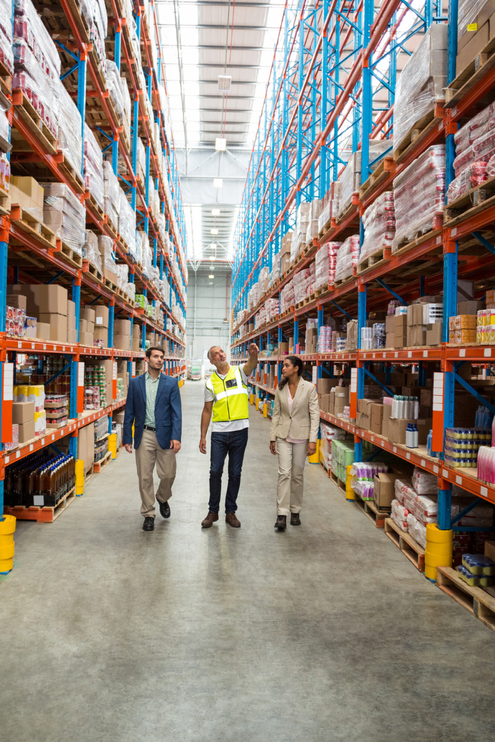 Three people are walking in a warehouse filled with pallets wrapped in plastic. The person in the middle is wearing a yellow vest and is pointing to the pallets.  The two people next to him are wearing business clothes.