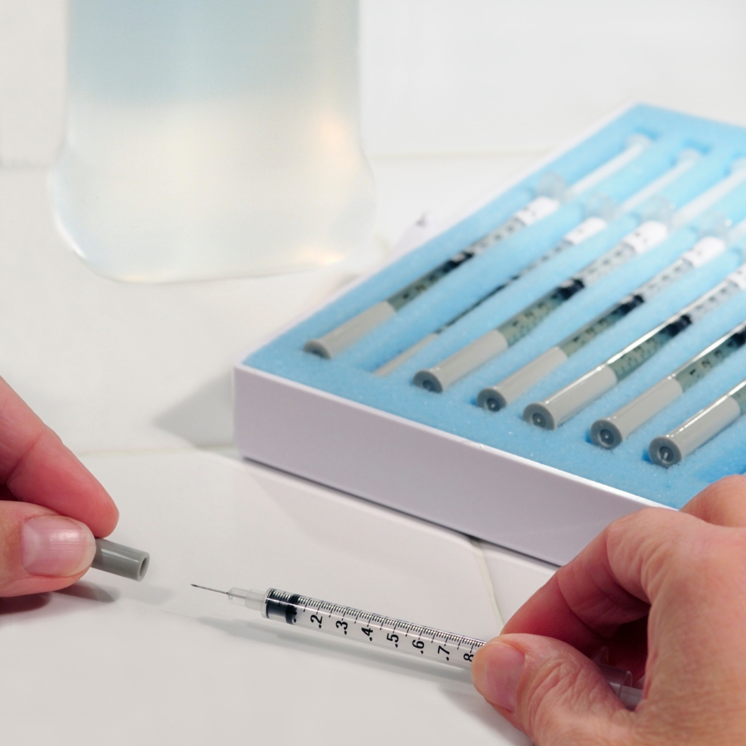 A person is holding an injection needle in front of a box containing a cardboard layer and foam holding various needles inside.