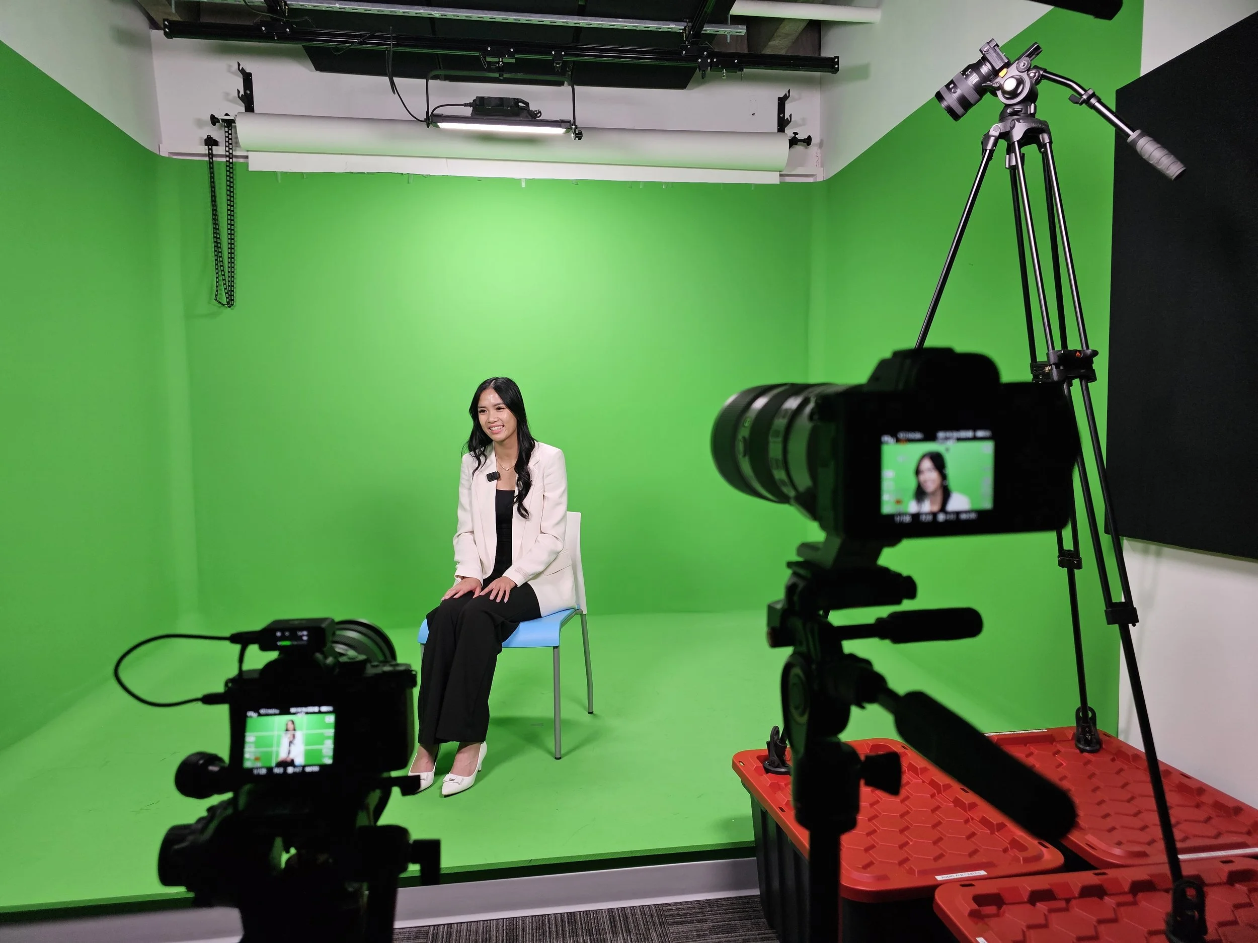 A woman seated in a recording studio with a green screen background, two cameras pointed at her, one on a tripod and the other on a tripod, capturing her image for a video or interview.