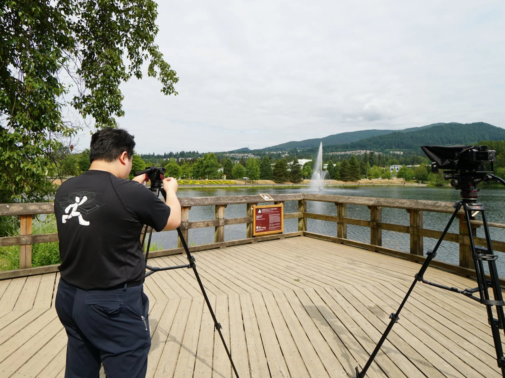 A person operating a camera on a tripod by a lake with a fountain and mountains in the background.