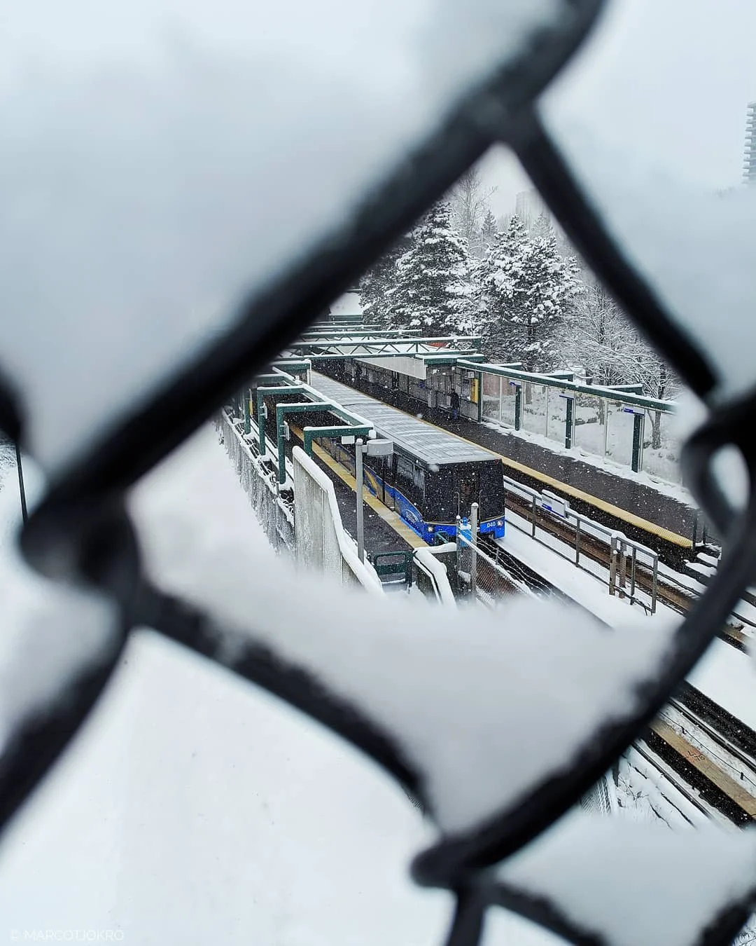 Looking through a window with snow on the glass, a blue train on a snowy train station platform with trees covered in snow in the background.