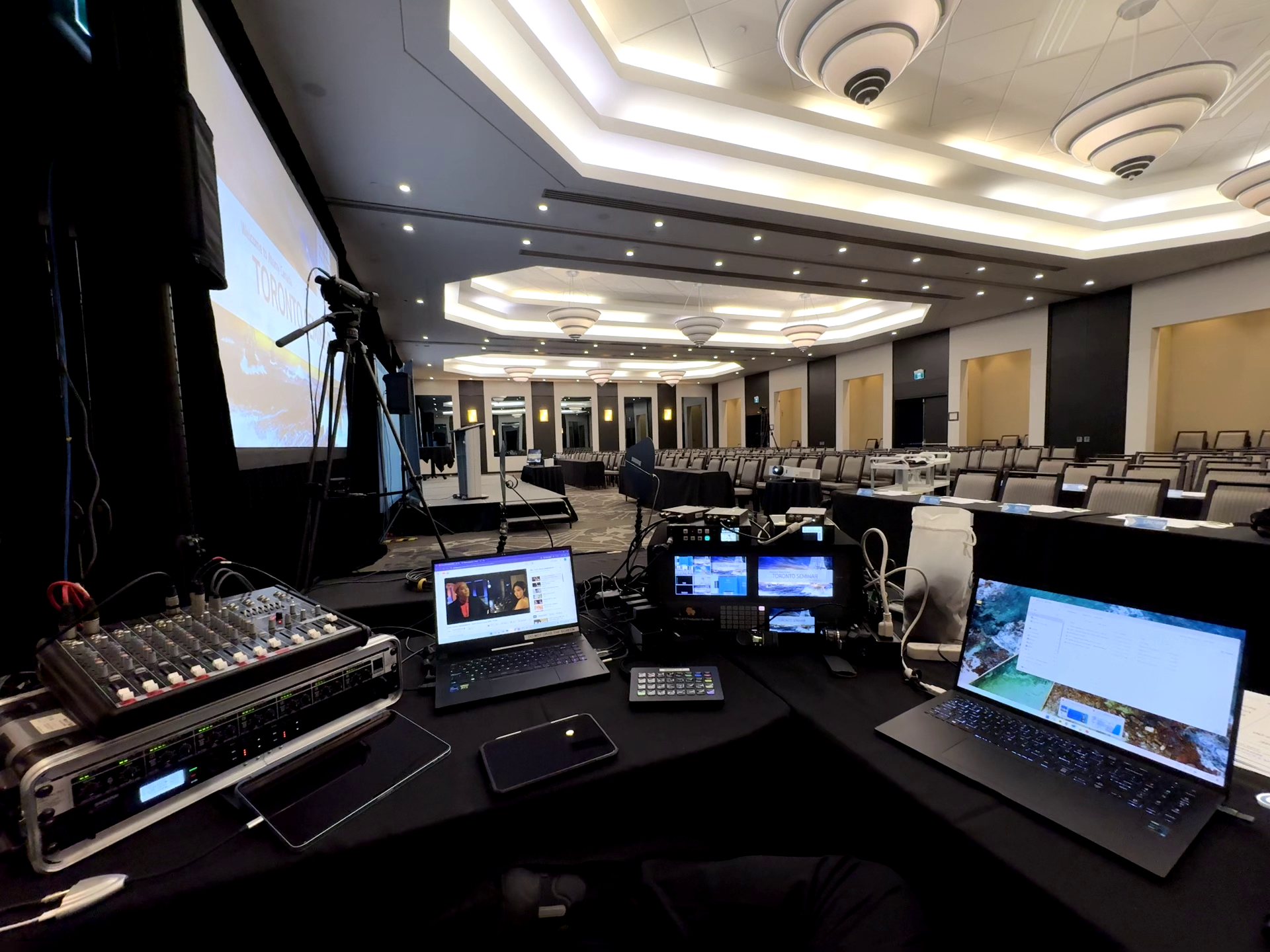 View of a conference room with empty chairs and a stage set up for a presentation, with audio-visual equipment and cameras in the foreground.