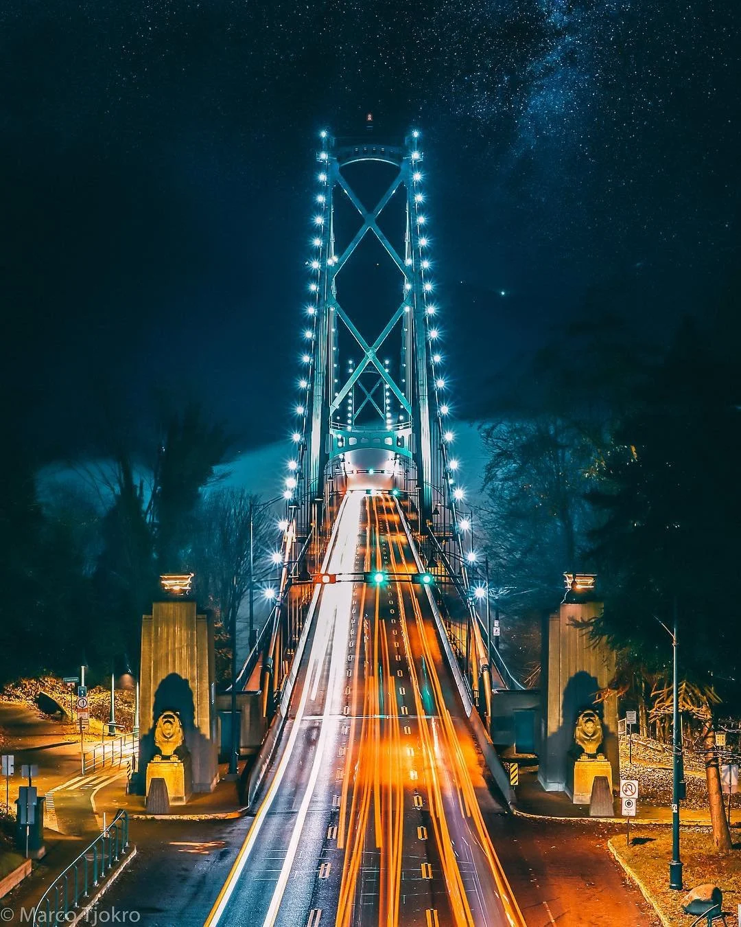 Nighttime view of a bridge illuminated with bright lights, showing light trails of moving vehicles crossing over, with a starry sky visible above.