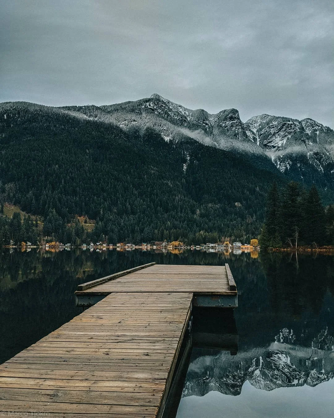 A wooden dock extends into a calm lake with a mountain range and forested landscape in the background.