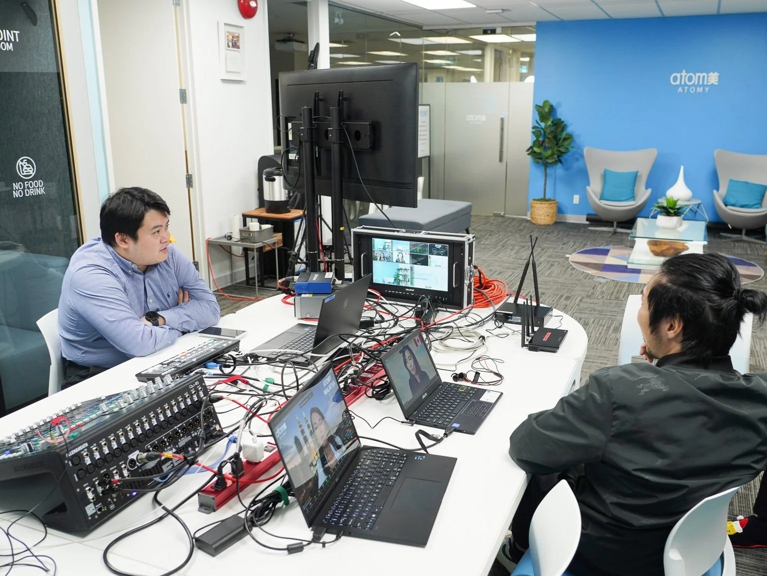 Two people are working at a conference table with multiple laptops, monitors, and cables. They are seated across from each other, in a modern office with a blue and gray lounge area in the background, featuring chairs, a small table, and plants.