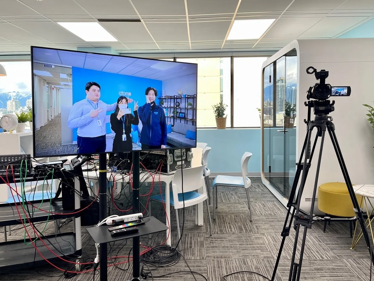 An office with a large monitor displaying three people taking a selfie, a camera on a tripod, and wired electronic equipment on a table. There are chairs, potted plants, and windows with a city skyline in the background.