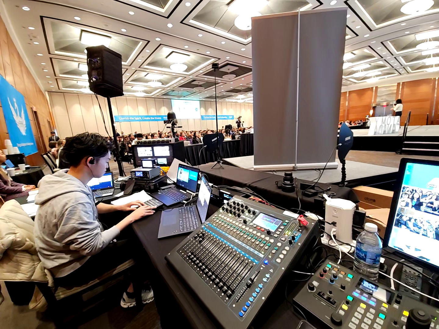 A conference or event hall with a stage and audience, a sound engineer operating equipment, and people seated in the audience.