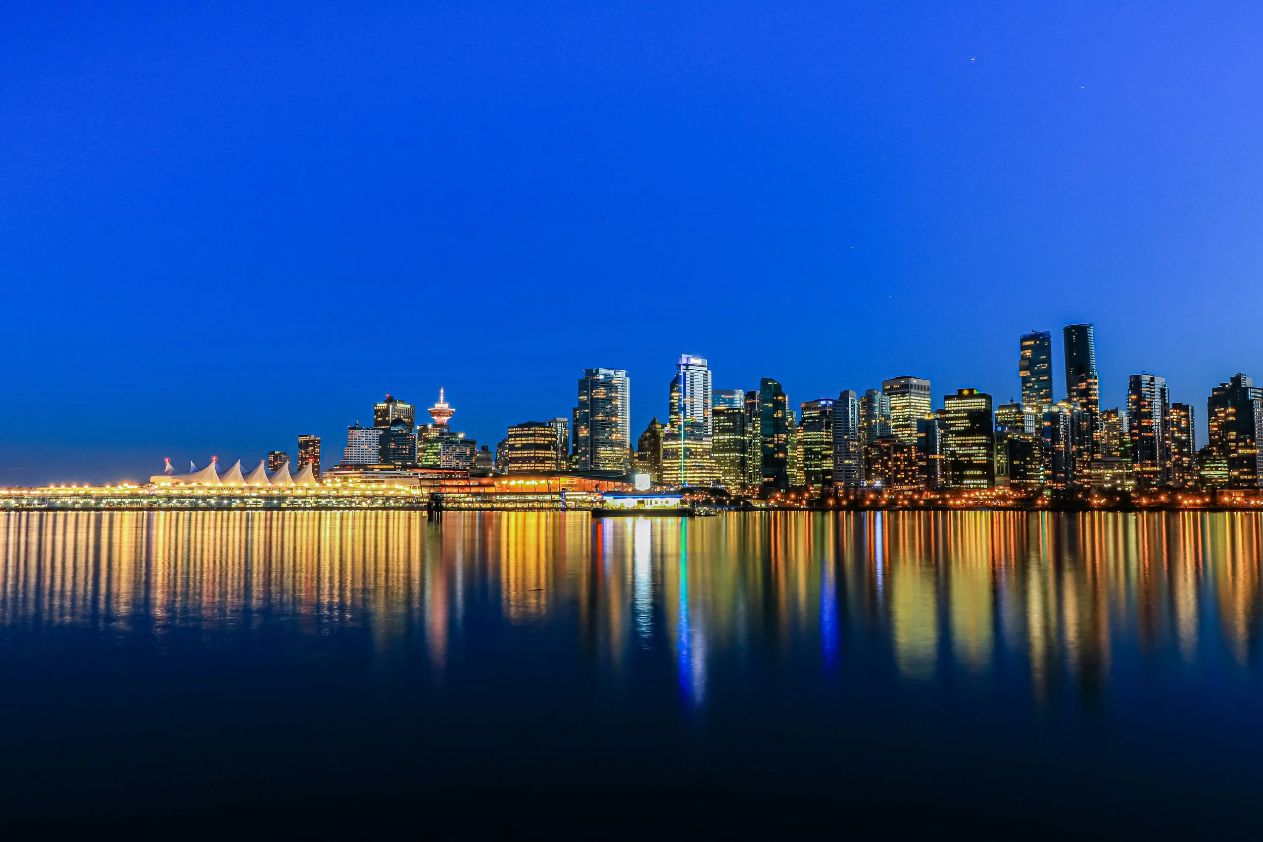 Night view of a city skyline with illuminated skyscrapers, reflecting on the water in front.
