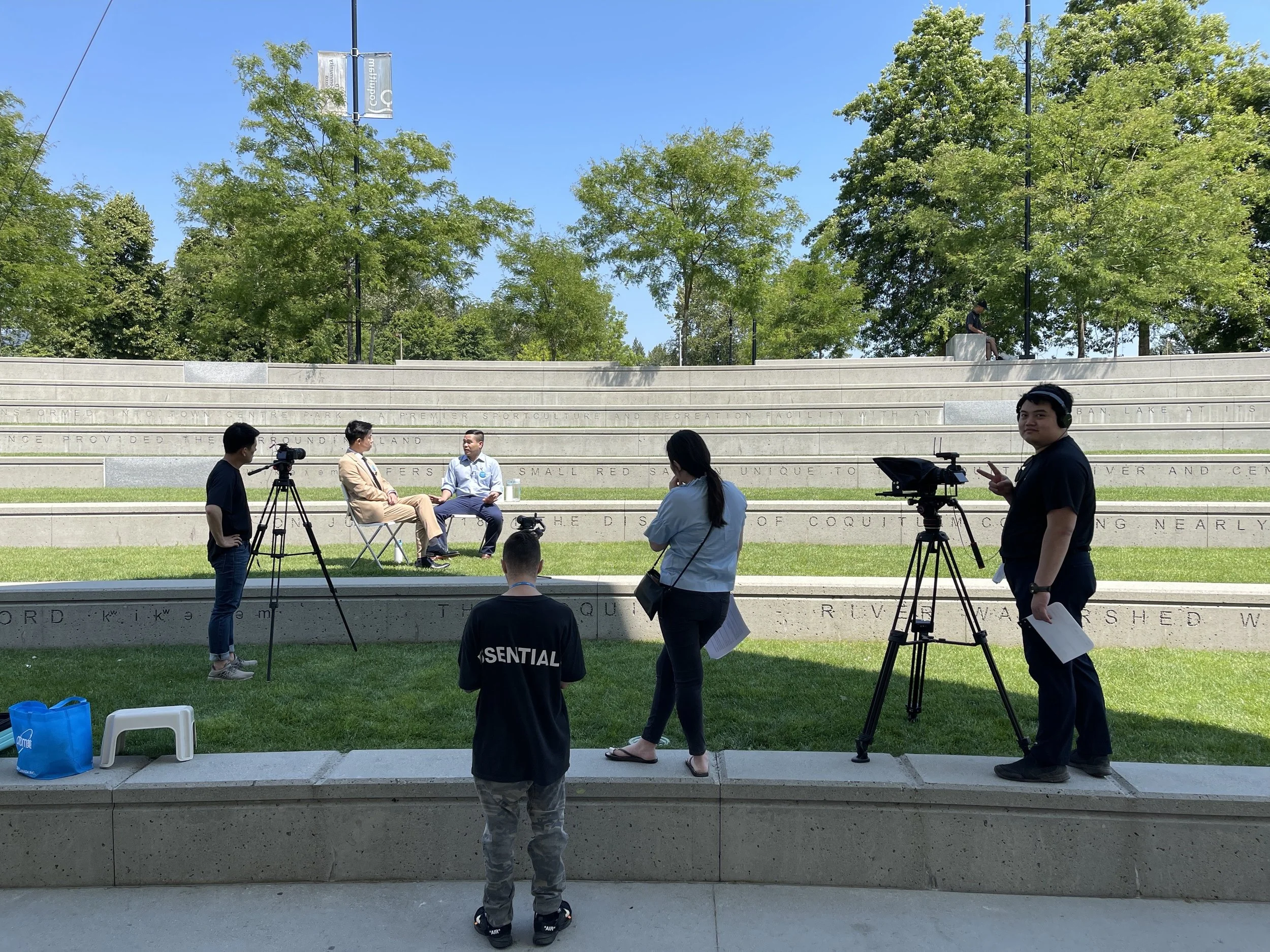 Filming of an interview or documentary in an outdoor park, with two men seated on chairs and crew members with cameras and equipment around them.