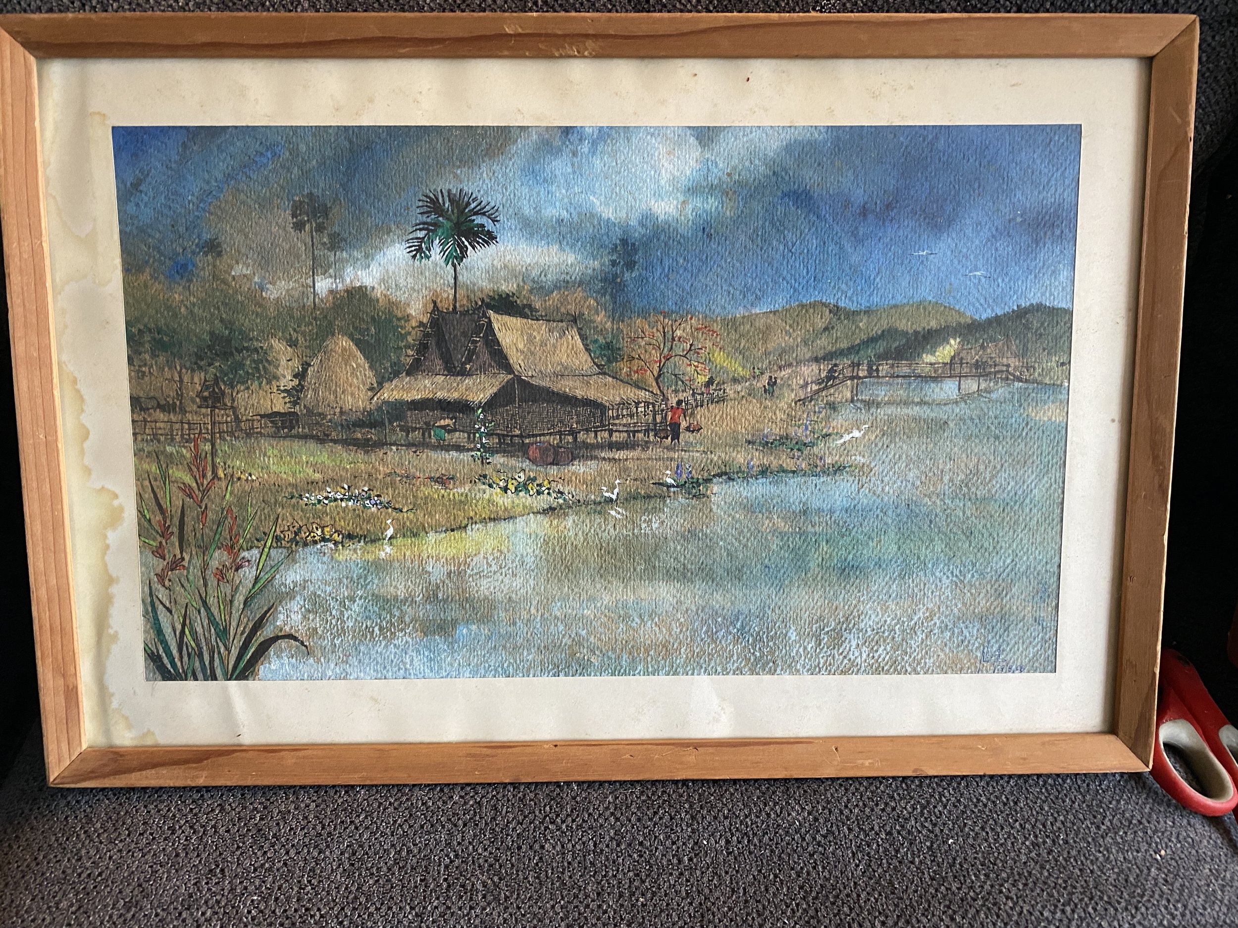 7) framed watercolor. 18.5x12.5". Indonesian palm leaf house on stilts facing river. Sig hard to read- Nouri or Lbuzi?  Dated (29/8/56 or 58) $500