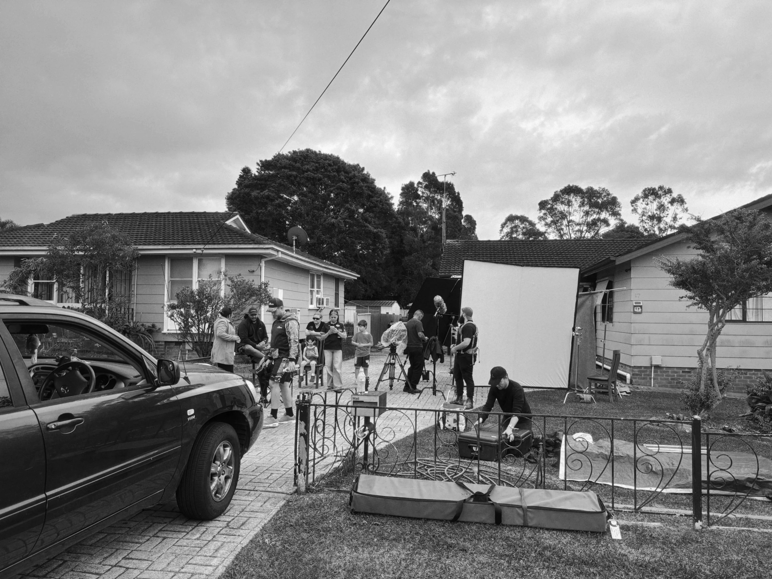 A group of people gathered outdoors in a residential neighborhood, preparing equipment for filming or photography. There are houses with tiled roofs behind them, and trees and cloudy sky in the background. A black car is parked in the foreground.