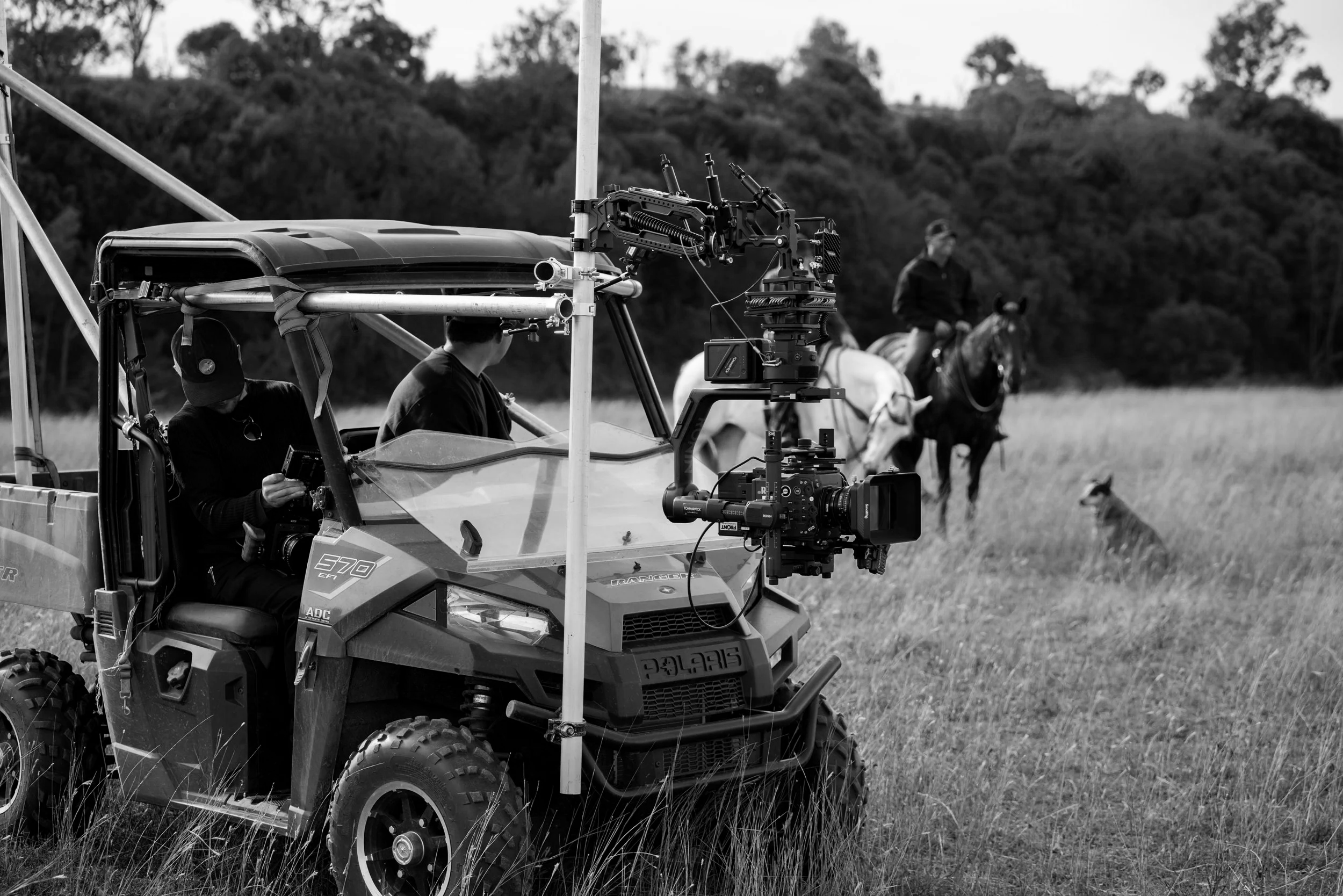 A film crew with a camera mounted on a robotic stabilizer on an all-terrain vehicle in a field. Two actors on horseback are in the background, with trees in the distance.