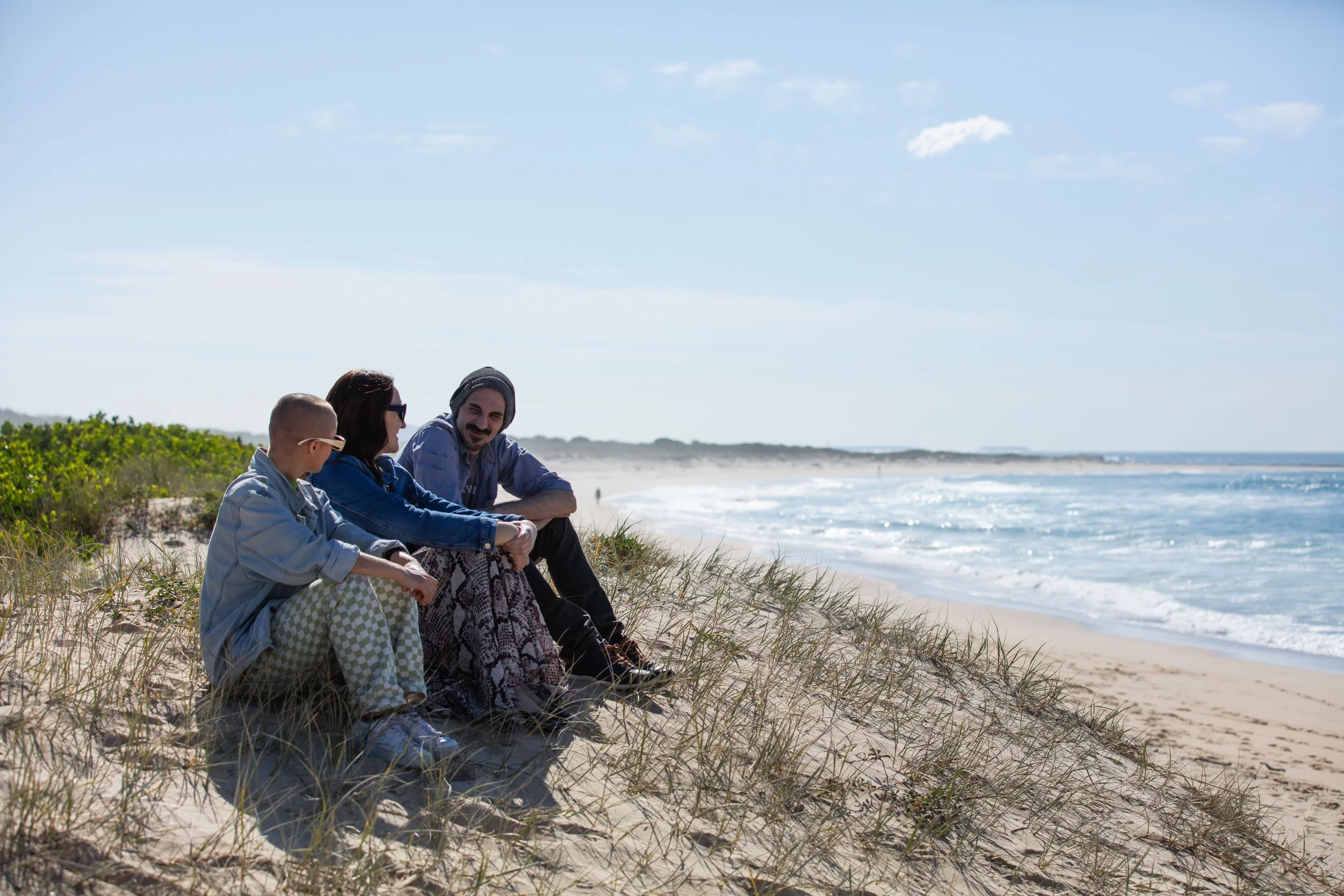 Four people sitting on the sand dunes at the beach, talking and enjoying a sunny day with the ocean in the background.