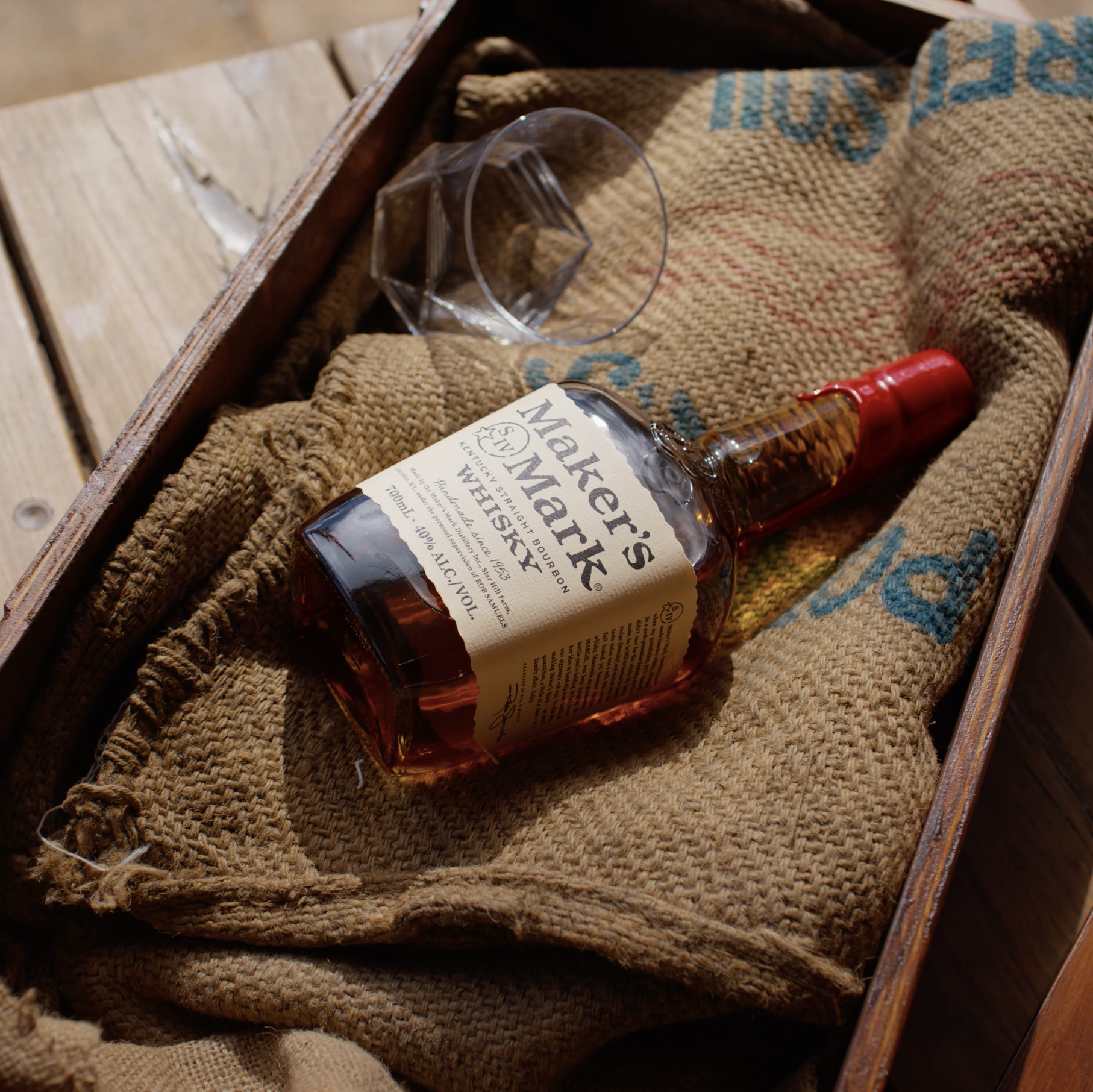 A bottle of Maker's Mark whiskey lying on a rustic wooden box lined with burlap fabric, with a glass tumbler nearby, on a wooden surface.