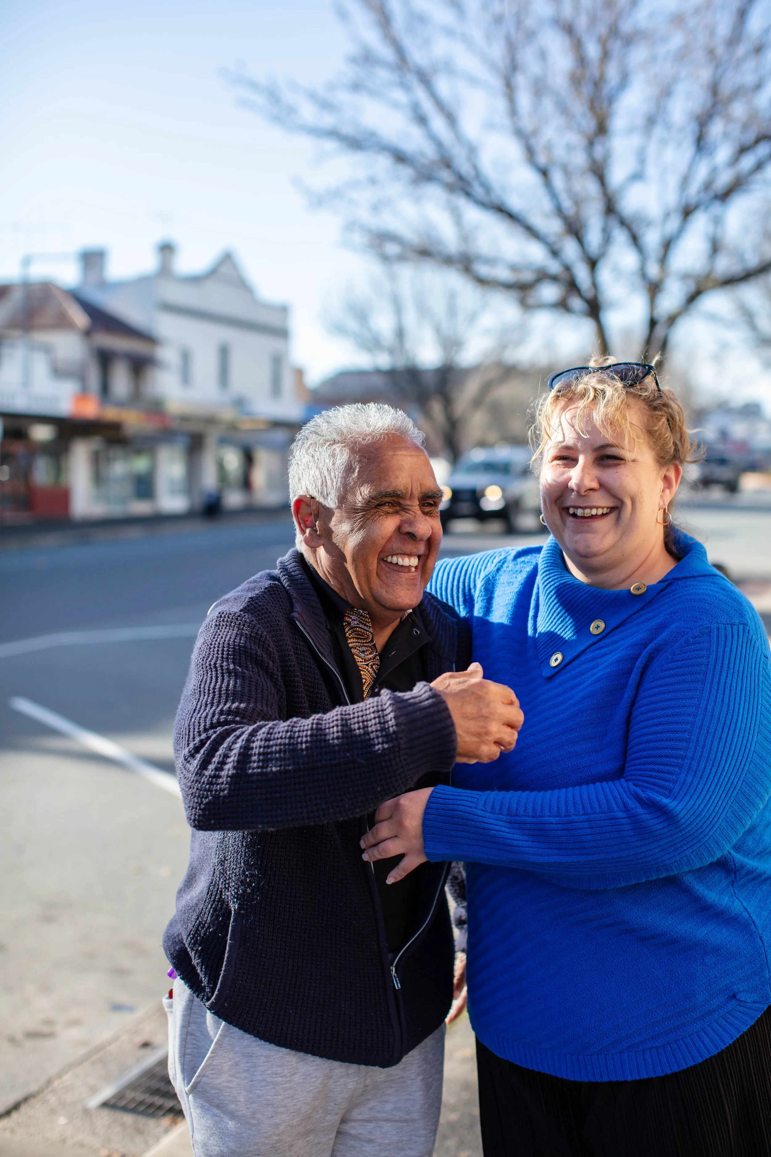 Two smiling women outdoors, hugging each other, with a background of leafless trees, buildings, and a street, on a sunny day.