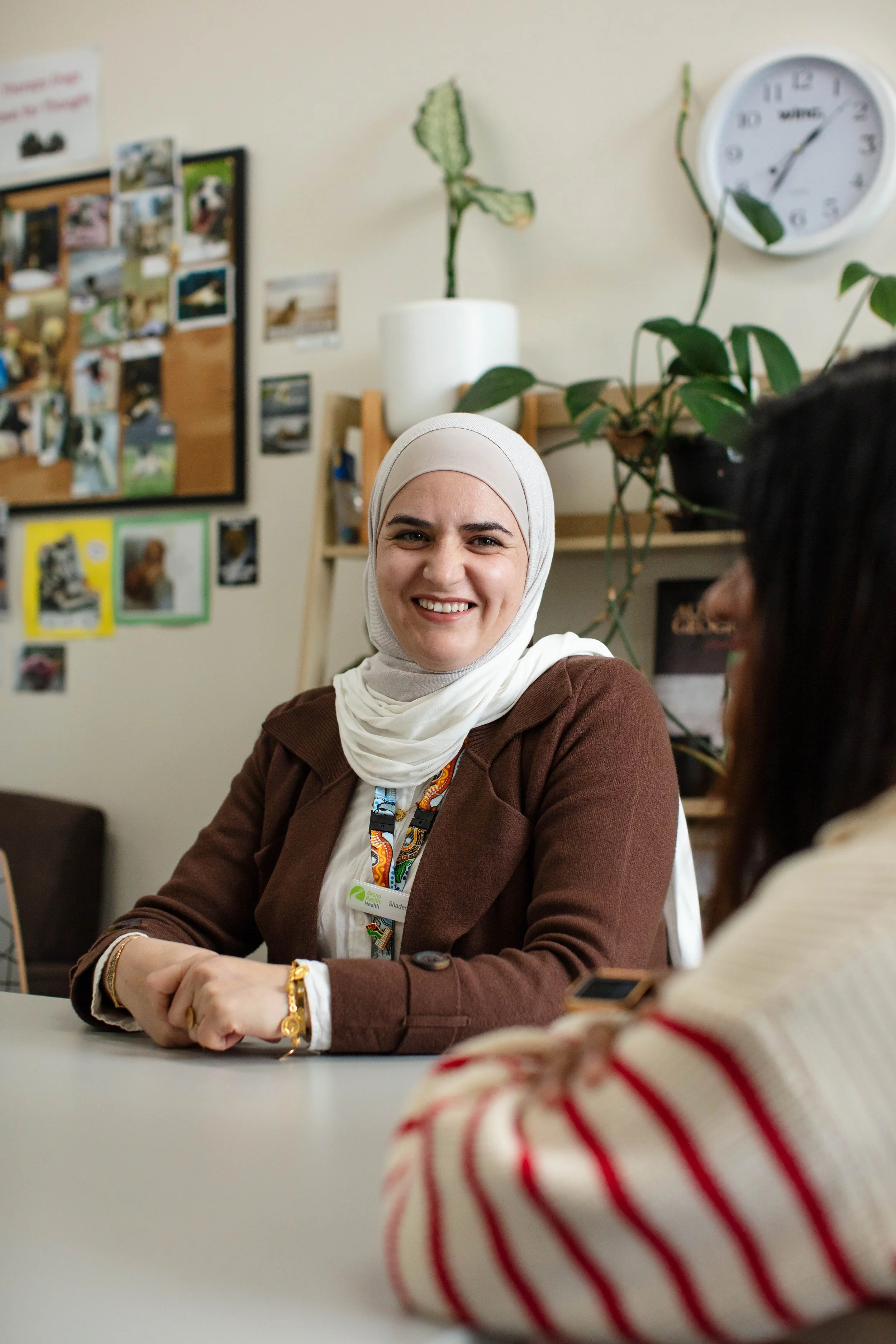A woman in a white hijab smiling during a meeting, with a blurred child in a red and white striped shirt in the foreground.