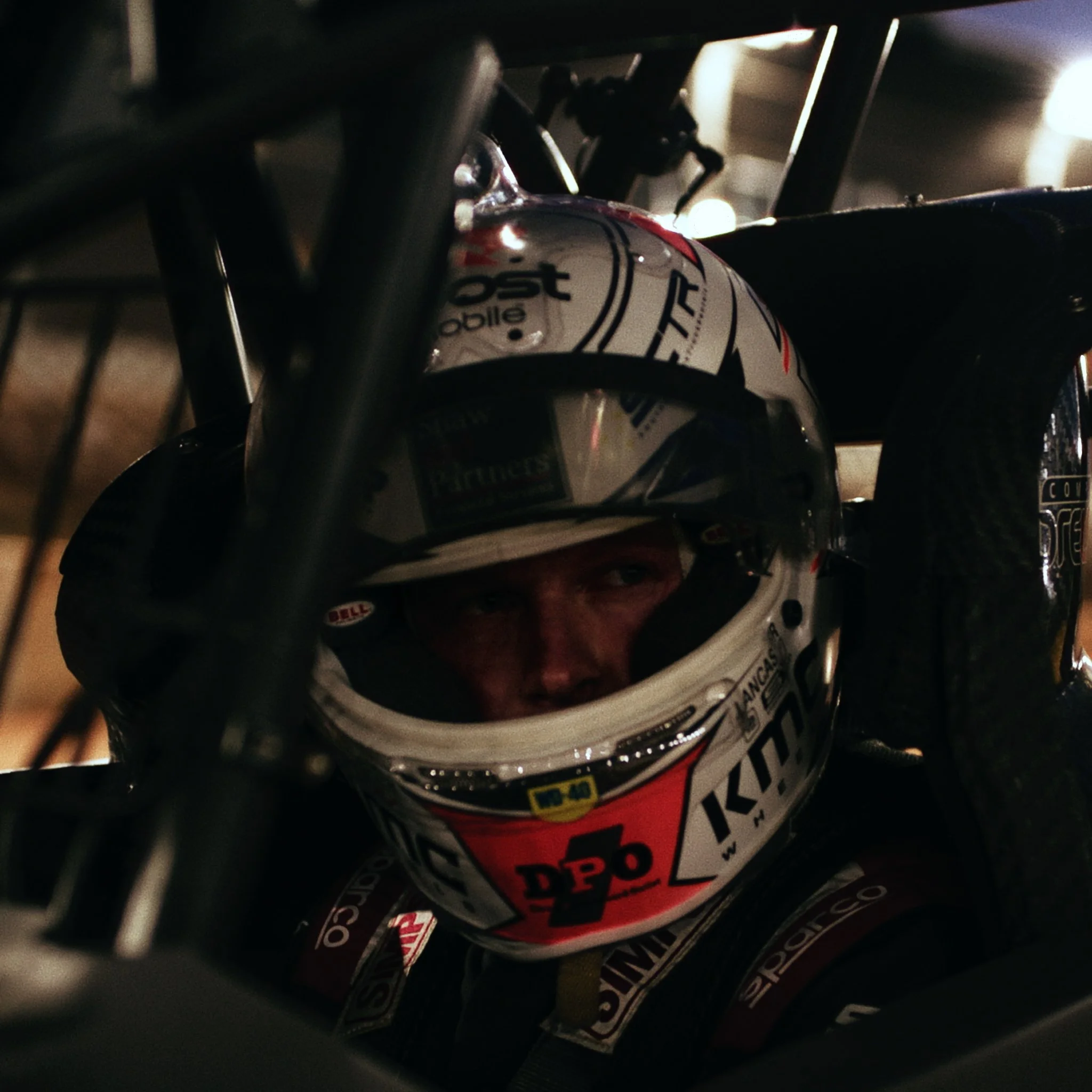 A close-up view of a racing helmet with sponsor decals, placed inside a vehicle, with parts of a roll cage visible in a dimly lit environment.