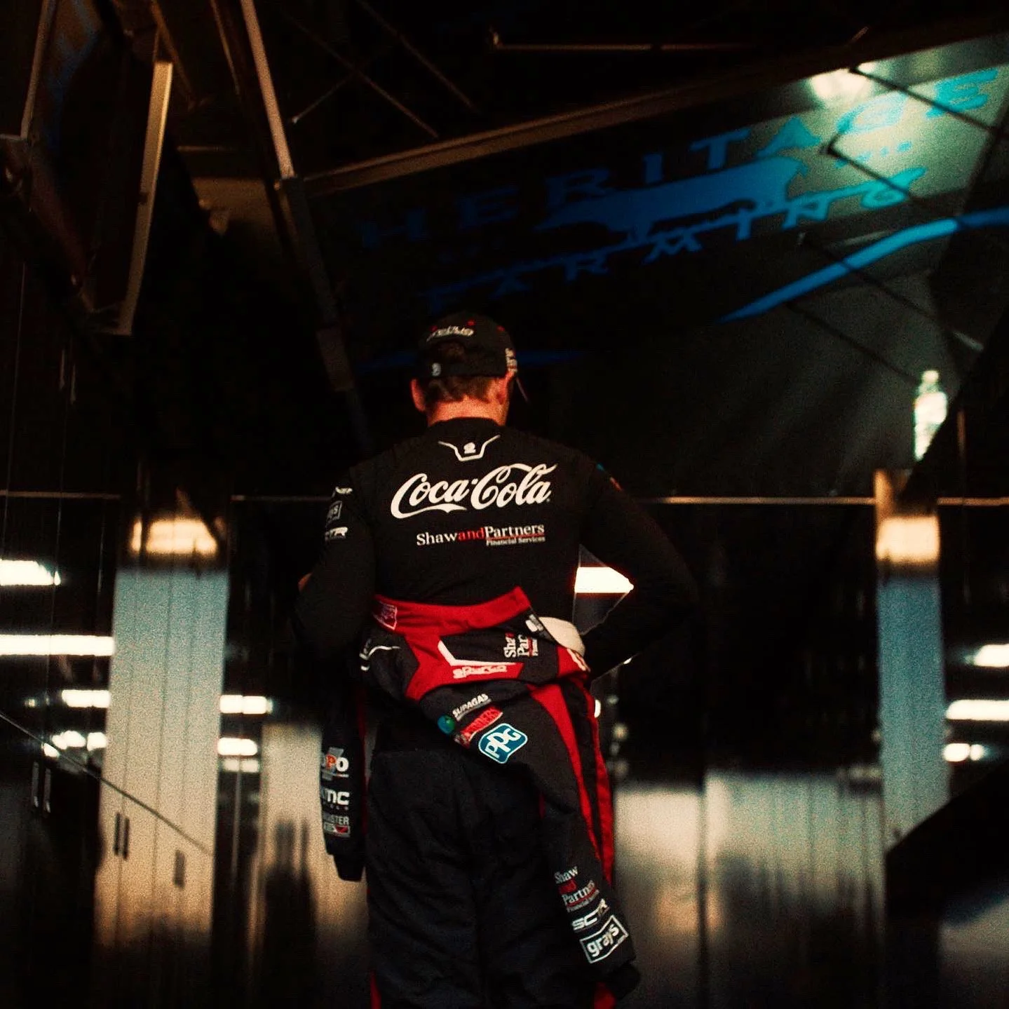 A person in racing gear with Coca-Cola and other sponsorship logos, standing in a dimly lit garage or paddock area, facing away from the camera.
