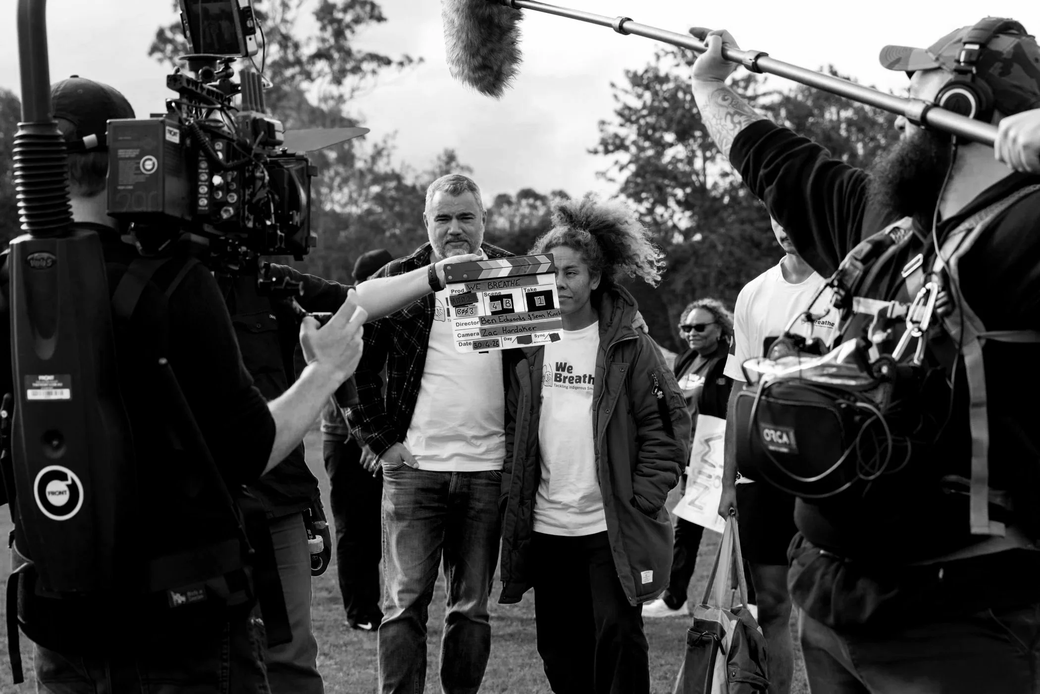 Black and white photo of a film crew filming a scene outdoors, showing a man with a beard and a curly-haired woman holding a clapperboard, with crew members and camera equipment visible.