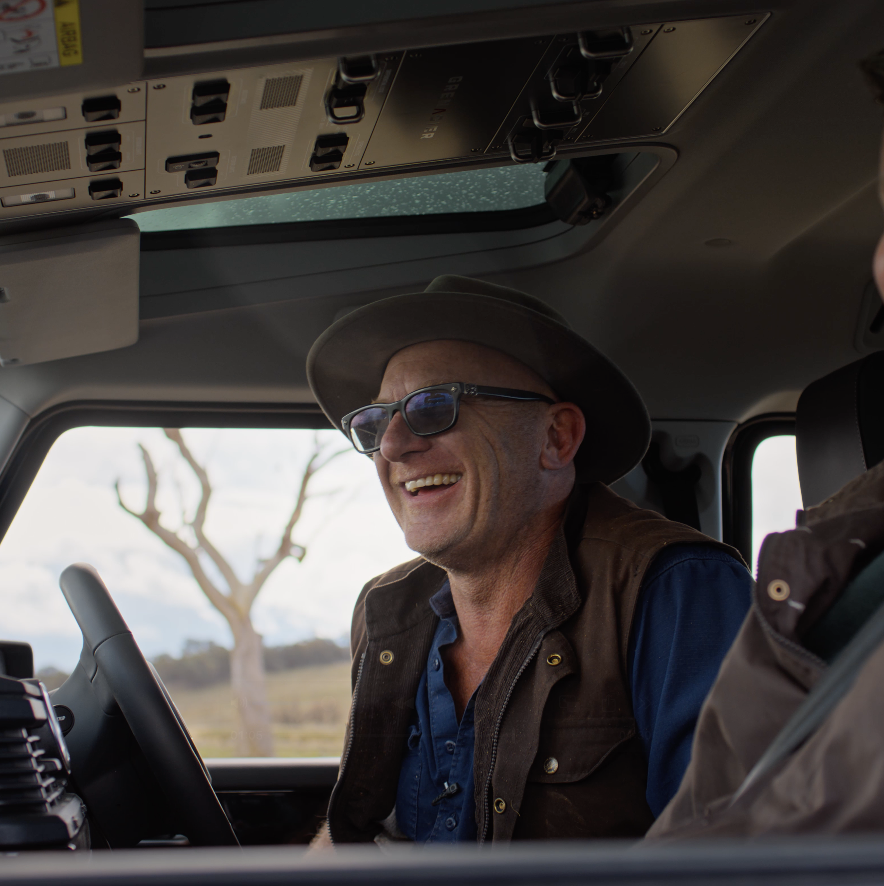 Man wearing glasses and a hat smiling while sitting in the driver's seat of a vehicle, with an open landscape and tree visible outside through the window.