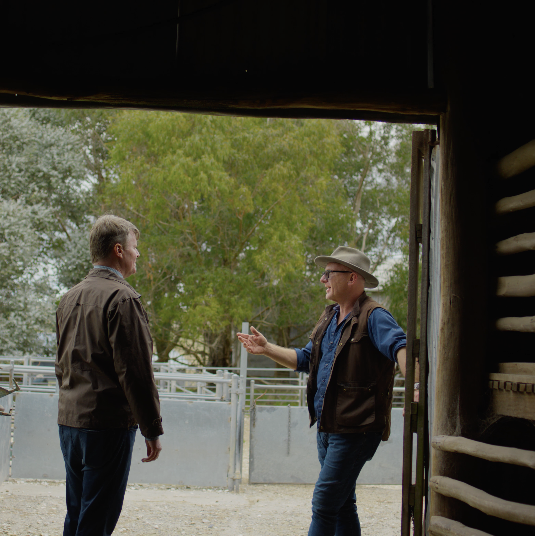 Two men engaged in discussion near a horse stable, with trees and fencing in the background.