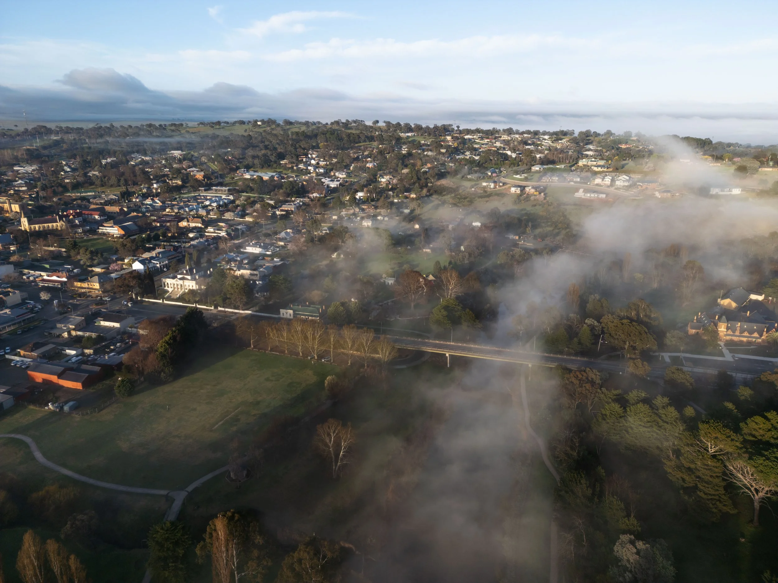 An aerial view of a suburban area with numerous houses, trees, green spaces, and a layer of fog or smoke partially obscuring the landscape.