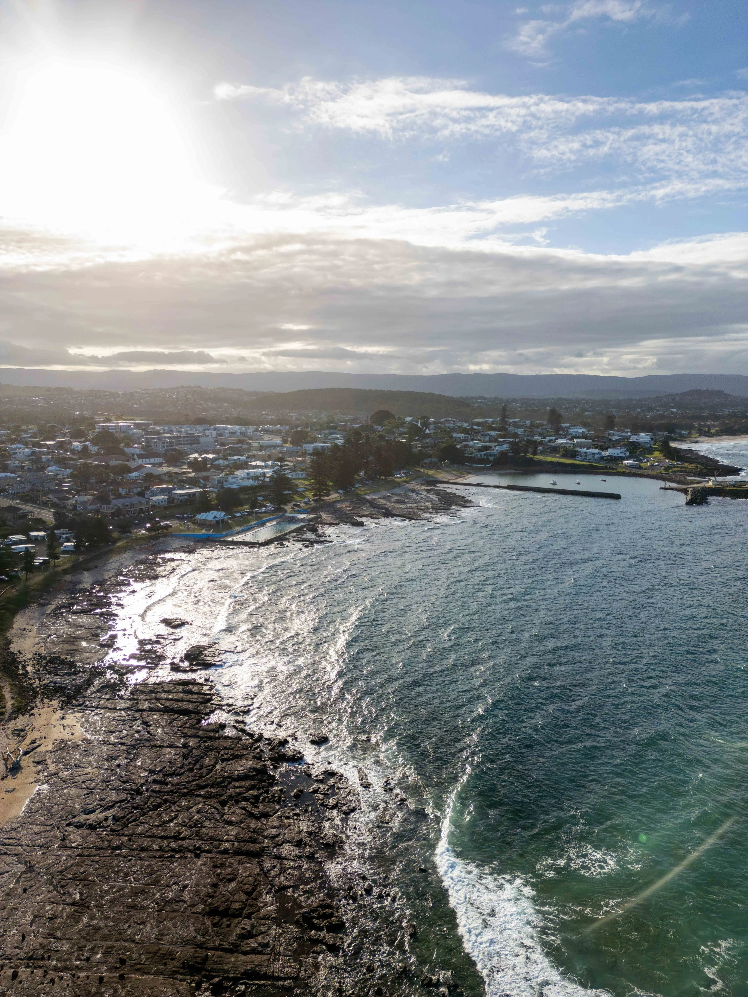 A coastline with rocky shore and waves crashing, a small harbor with boats, residential area, and hills in the background under a partly cloudy sky with the sun shining.