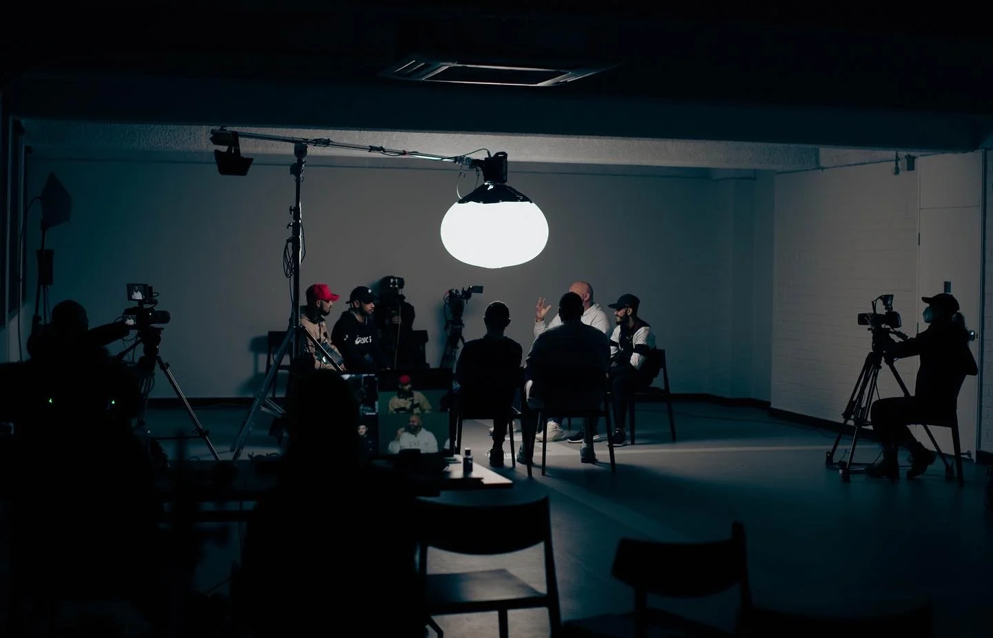 People sitting in a circle in a dark room with a large bright light overhead, cameras filming the scene, likely a recording or interview session.
