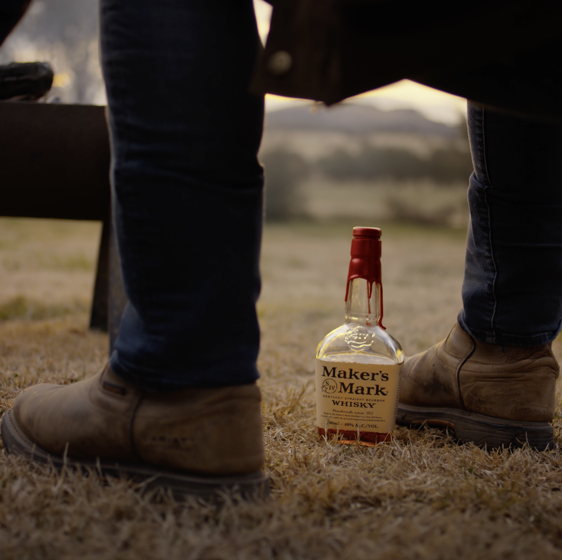 Close-up of two people's legs standing on dry grass near a bottle of Maker's Mark whiskey, with a bottle cap beside the whiskey bottle, and blurred background sunset or sunrise sky.
