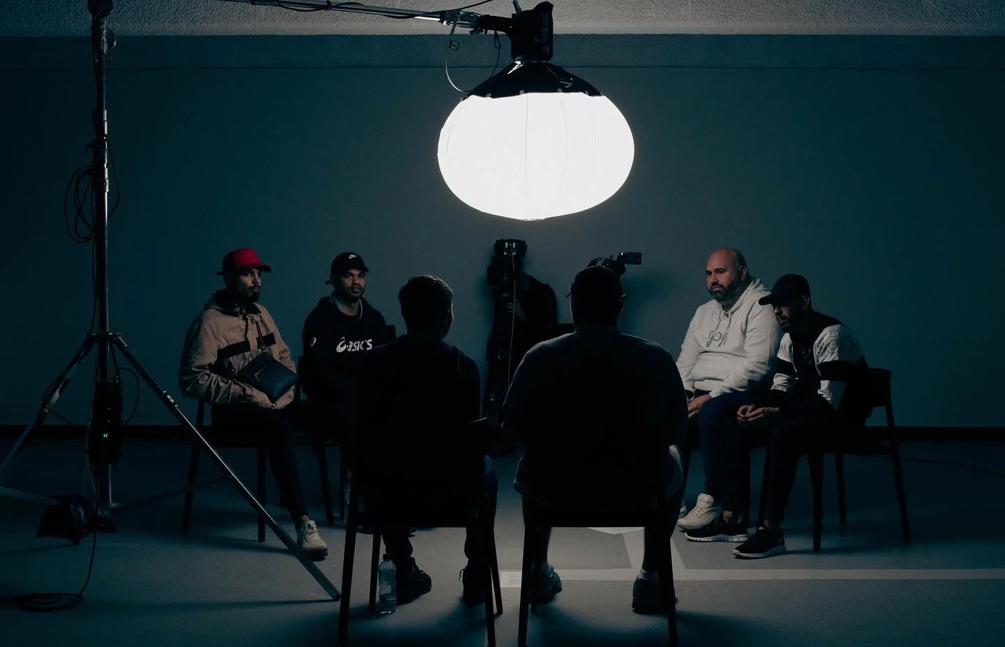 People sitting in a circle during a recording or interview session in a dimly lit studio with a large overhead light.