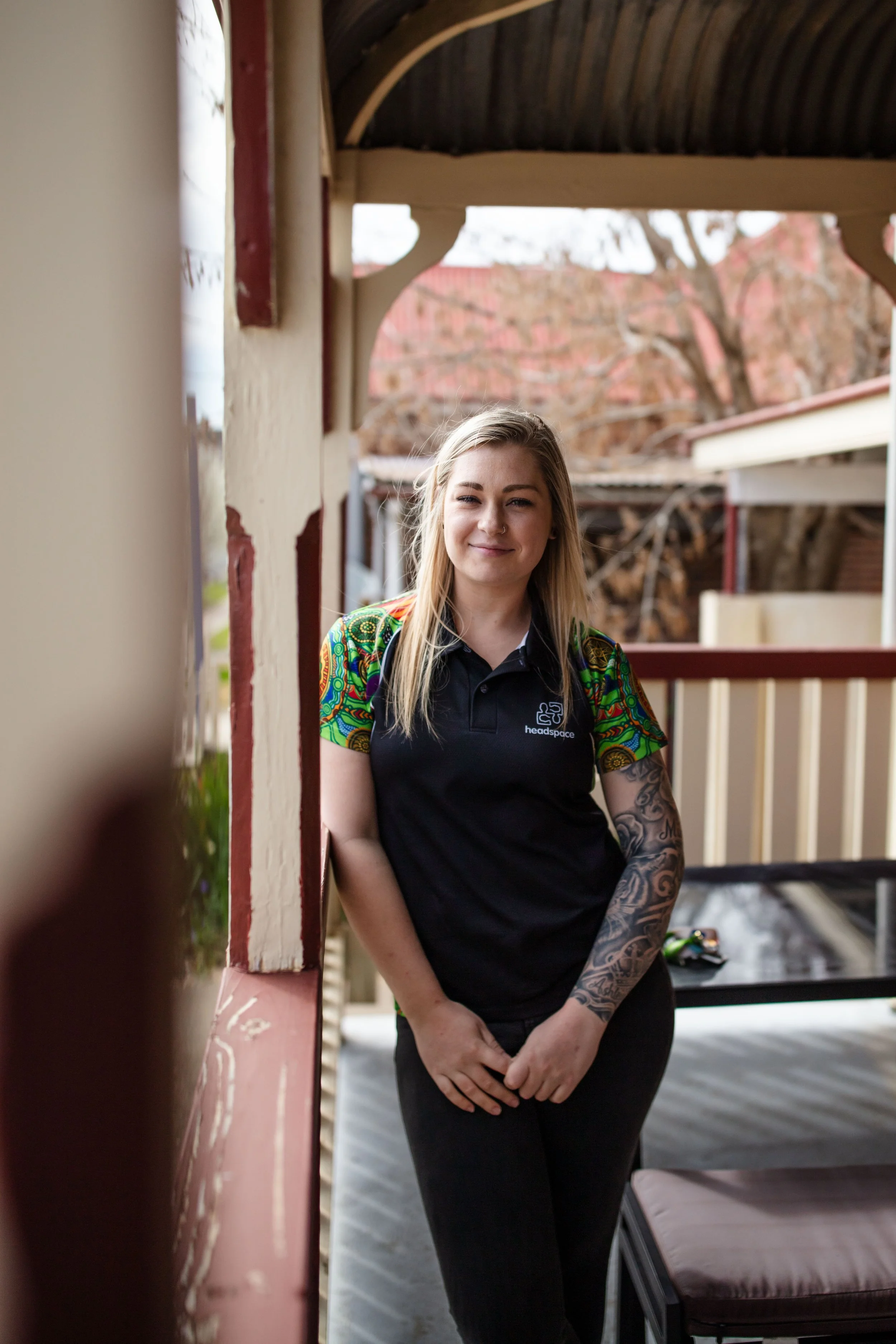 A young woman with long blonde hair and tattoos on her right arm, smiling softly while standing on a porch or balcony with a wooden roof. She is wearing a black polo shirt with a logo that reads 'headspace' and colorful sleeves. The background shows 