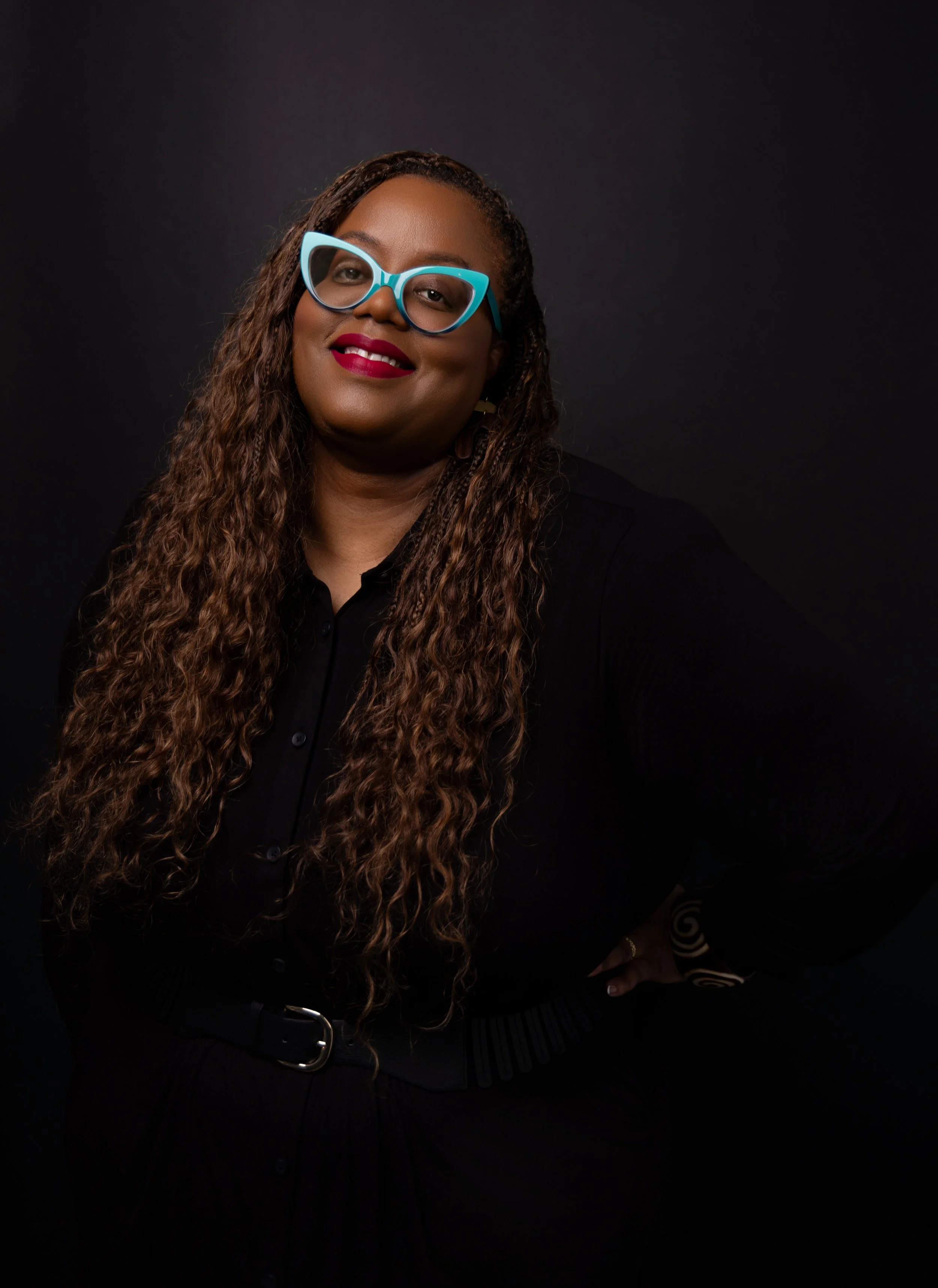A woman with long, curly hair wearing large, light blue glasses, red lipstick, and a black top, standing against a dark background.