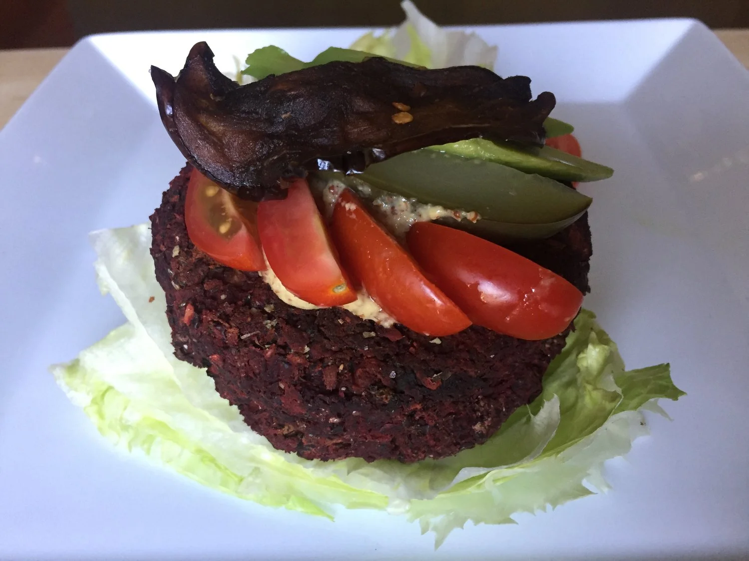 Beet, kidney bean and mushroom burger, on a lettuce bun, topped with a piece of eggplant bacon, pickles, tomato, and avocado.