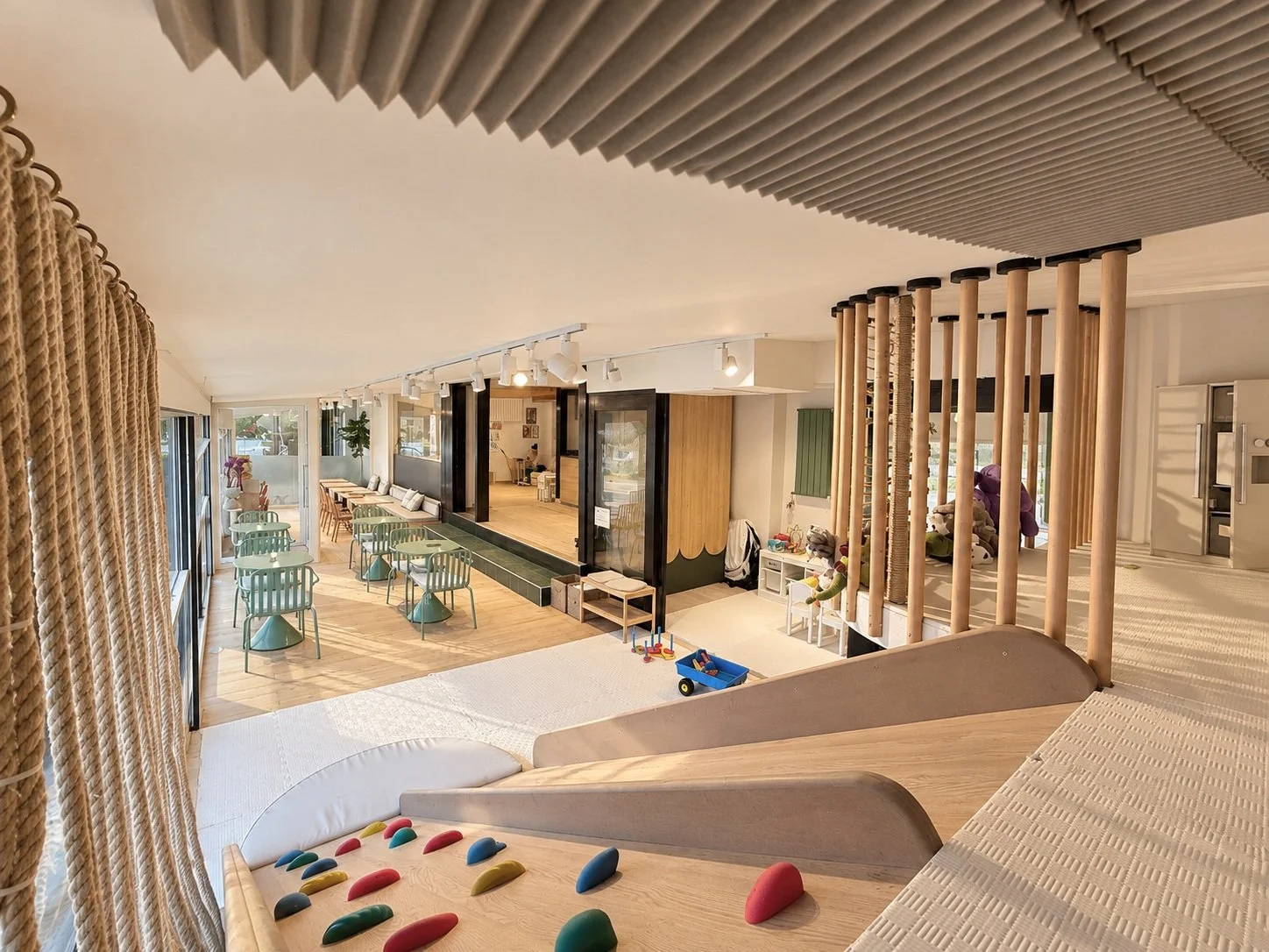 Children's indoor play area with a slide, climbing blocks, and toys. The space has large windows, a dining area with teal chairs, and a stage area with wooden flooring and lighting.