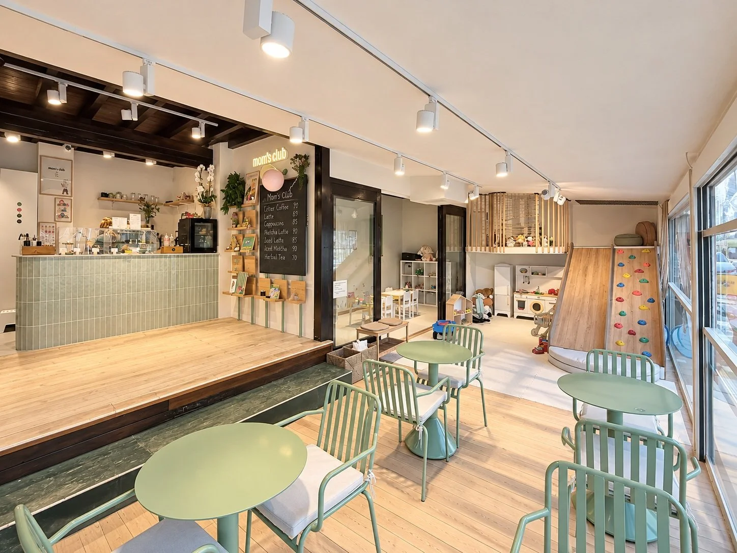Indoor space with pastel green tables and chairs, a serving counter, and a play area with a climbing wall and toys, bright large windows on one side