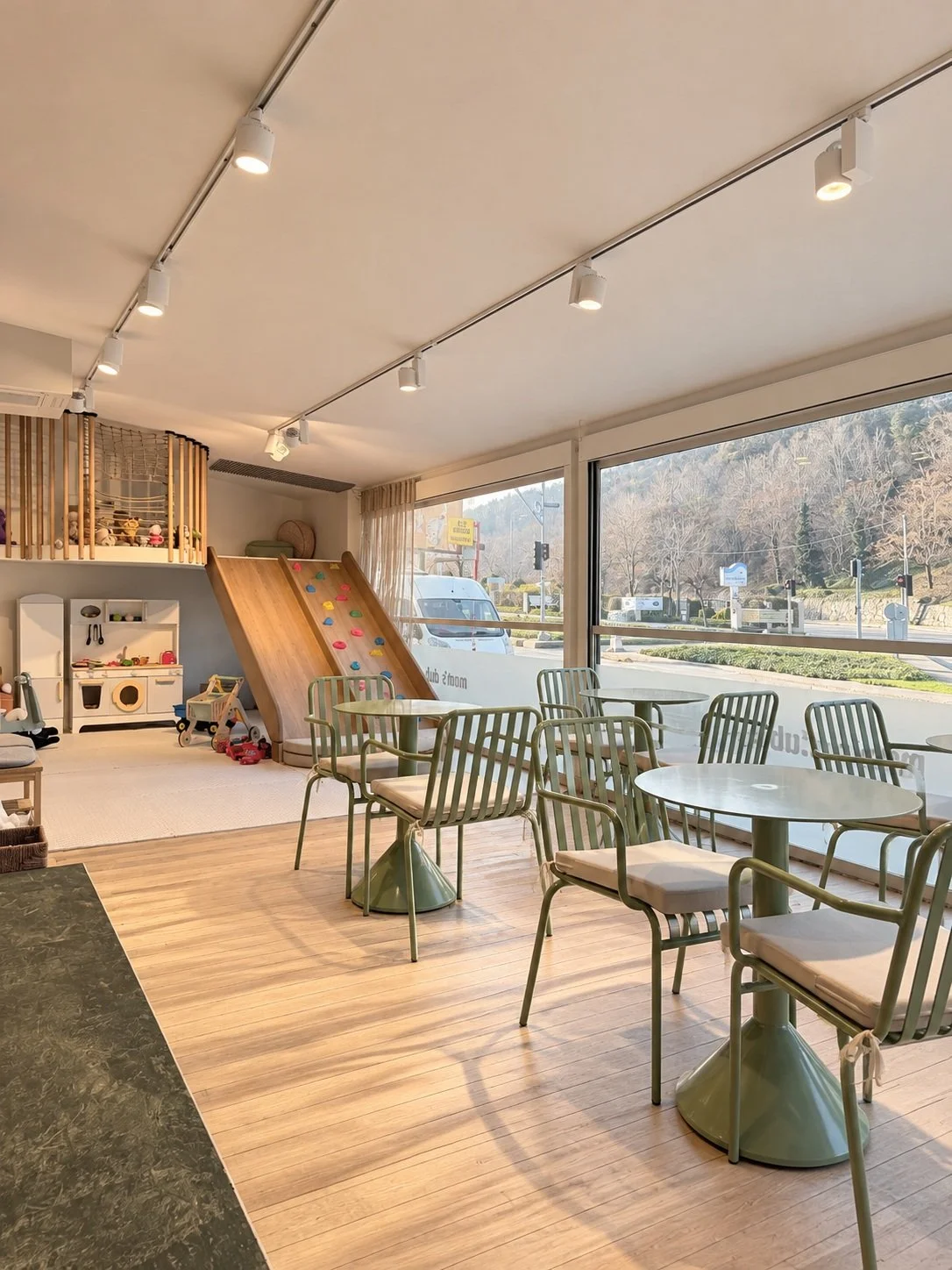 A cozy indoor children's play area with a wooden slide, colorful climbing wall, and toys in the corner, adjacent to a seating area with round tables and green chairs, large windows showing a street with trees and vehicles outside.