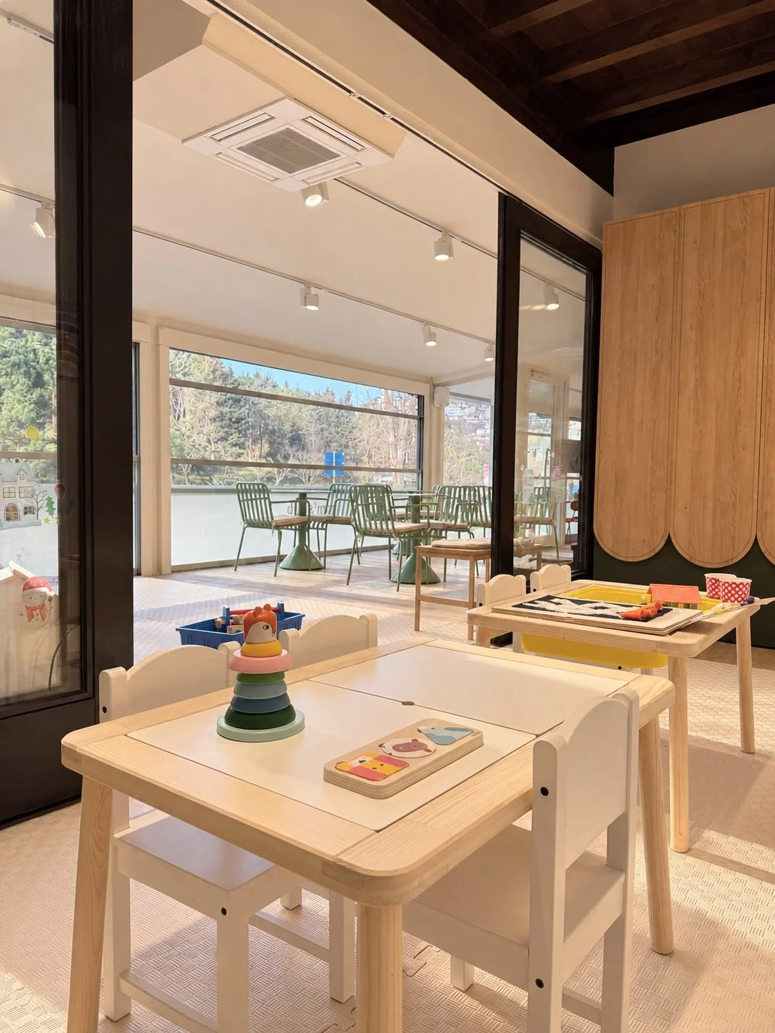 Child-sized white tables and chairs in a bright, modern indoor space with large windows and an outdoor balcony, decorated with toys and children's books.