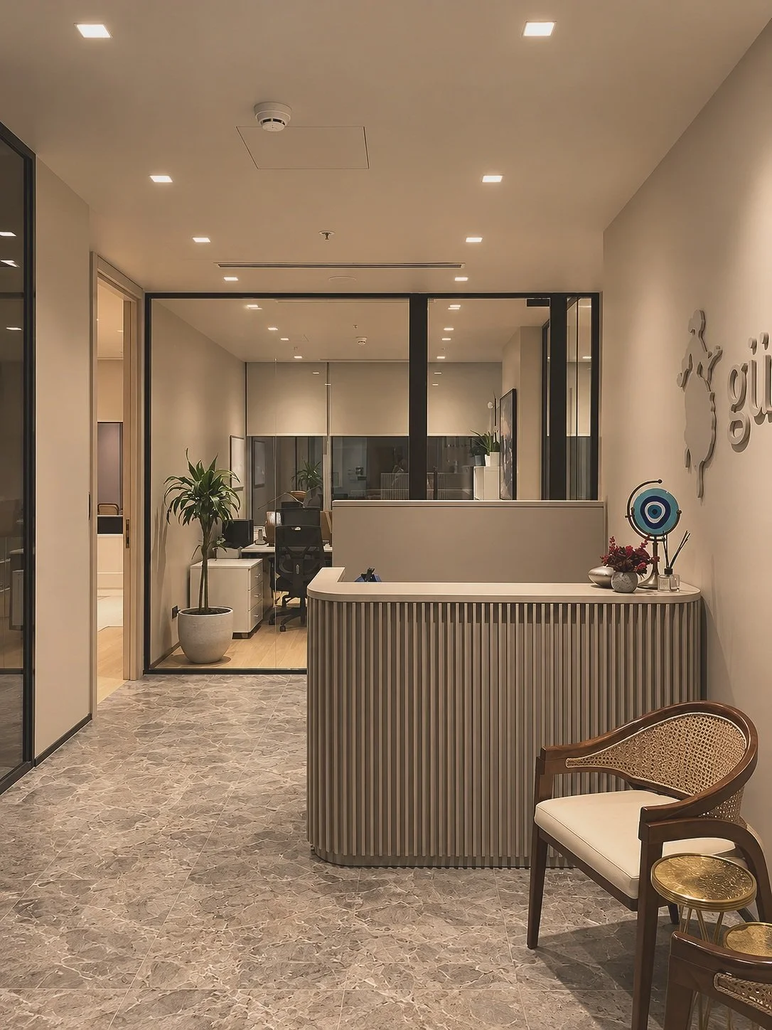 Modern office reception area with marble flooring, a curved reception desk, and a wooden chair with woven seating. Behind the desk, there's a wall with decor and a small round table with flowers and a decorative item. In the background, a glass partition separates the reception from the work area with plants and office chairs.