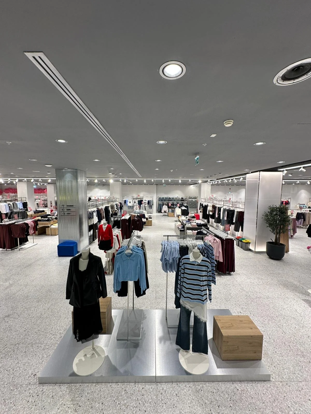 Empty clothing store with mannequins wearing sweaters and pants, racks of clothing, and a potted plant in the background.