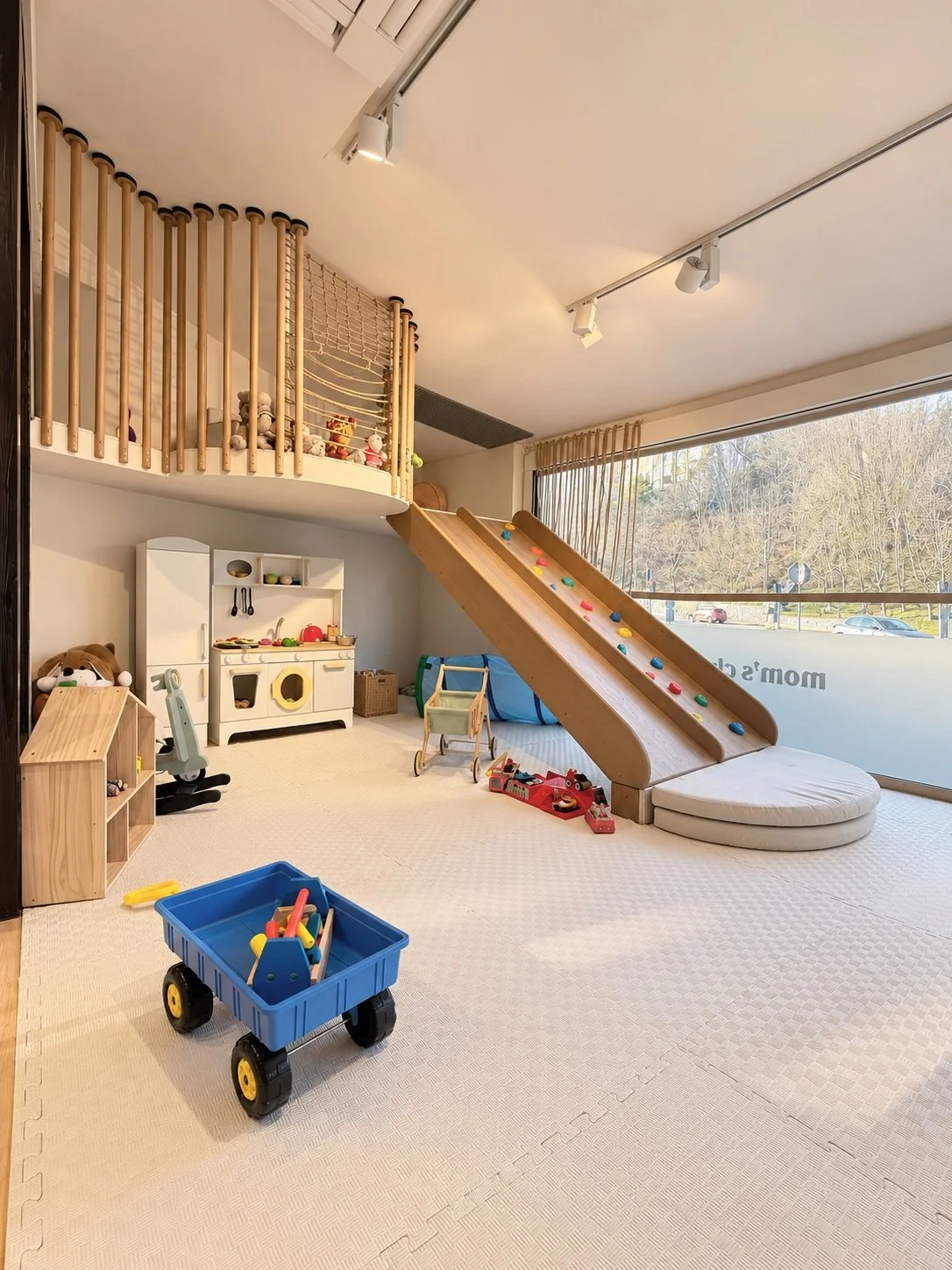 Indoor children's playroom with a wooden slide and climbing wall, toy kitchen, stuffed animals, and play equipment, near large window showing trees outside.
