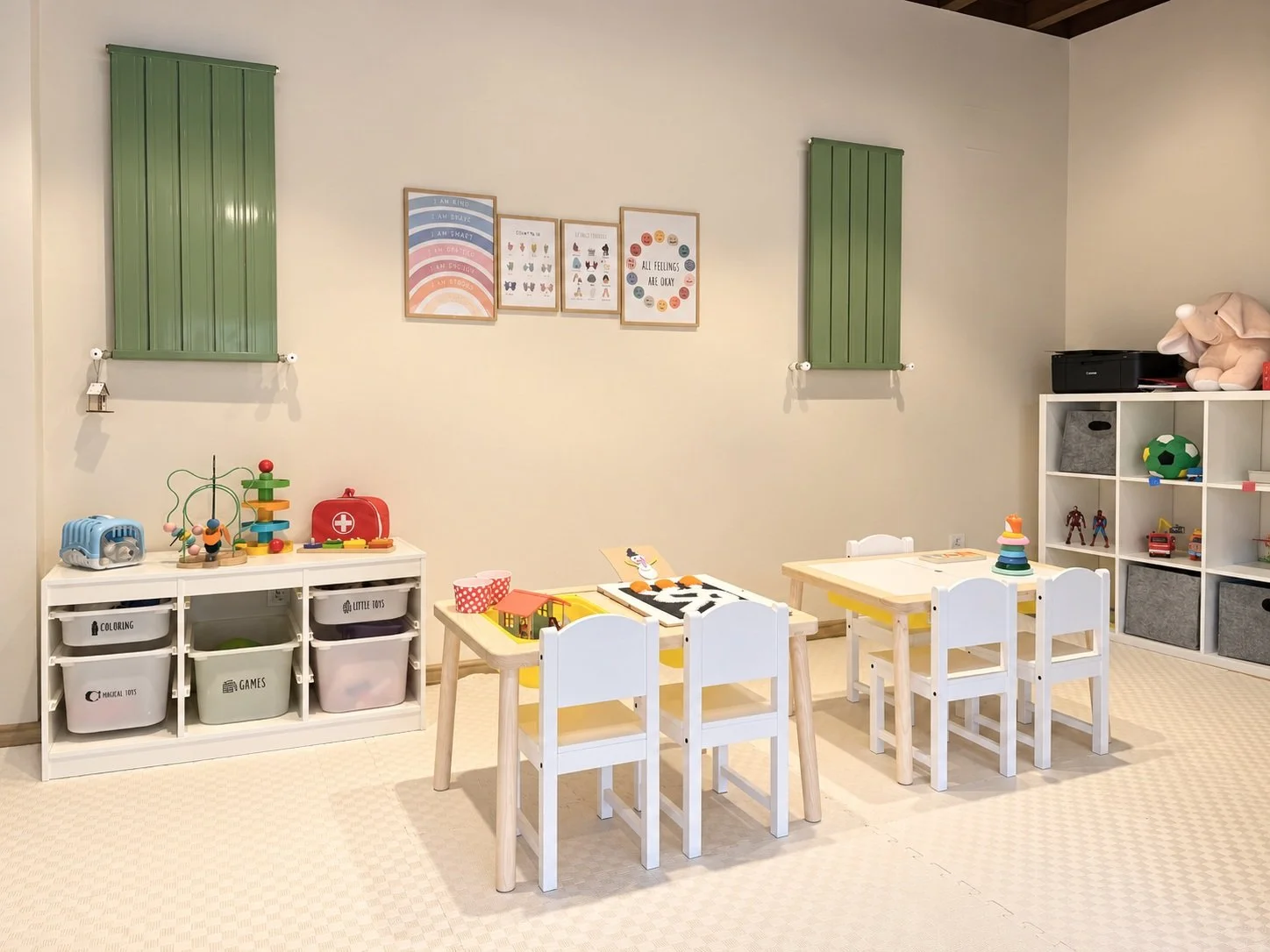 A children's playroom with small white tables and chairs, shelves with toys and storage bins, wall art, and green radiator covers.