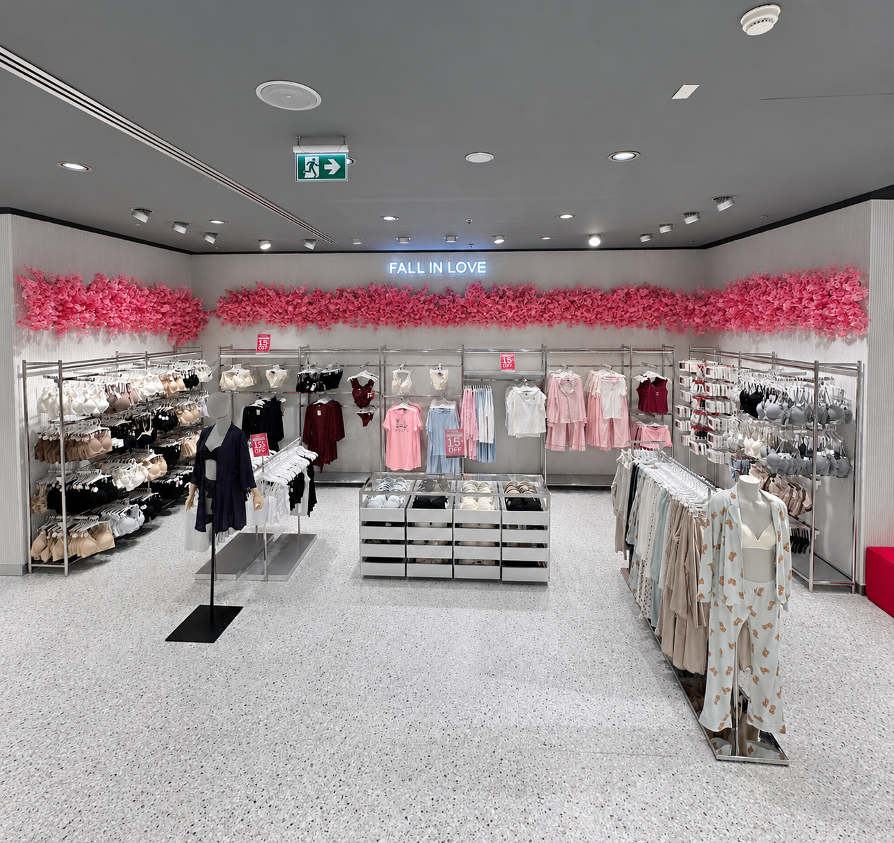 A lingerie store display with various bras, underwear, and pajamas, decorated with pink floral accents and a sign that says 'Fall in Love'.