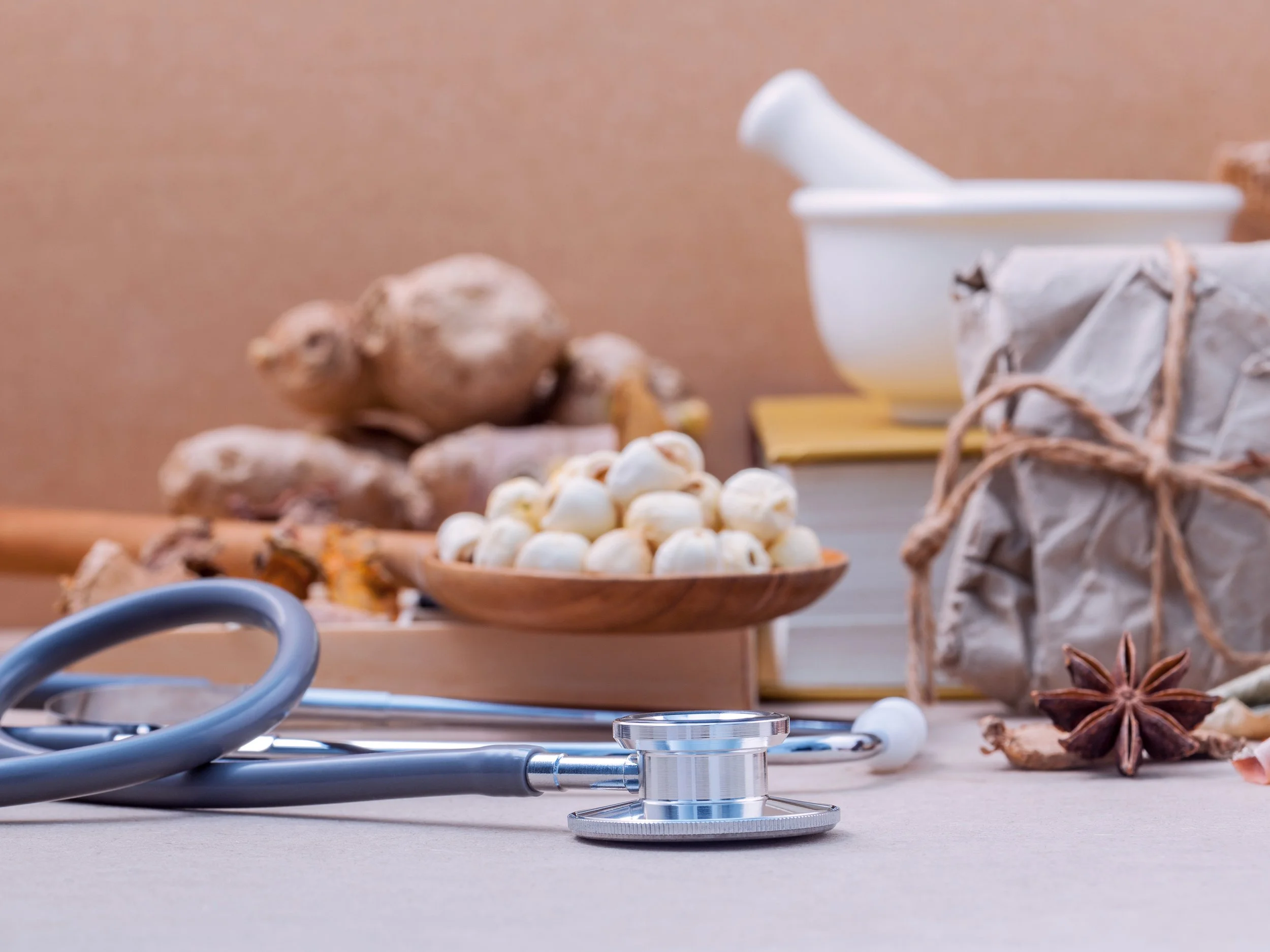 Medical stethoscope placed on a beige surface with herbal ingredients like ginger, garlic, star anise, and a mortar and pestle in the background.