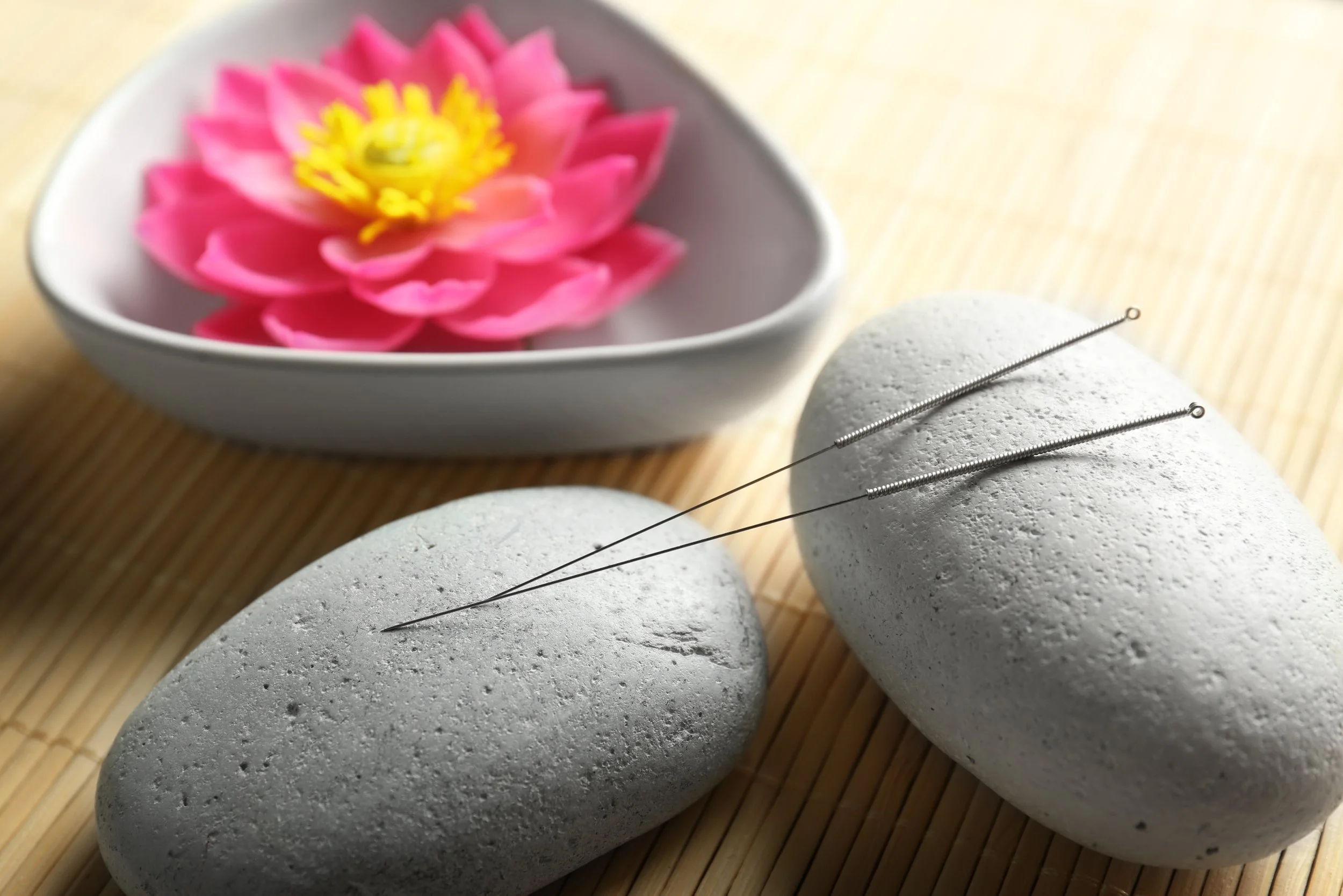Two smooth white stones with acupuncture needles placed on them, a pink lotus flower in a white bowl, all on a bamboo mat.