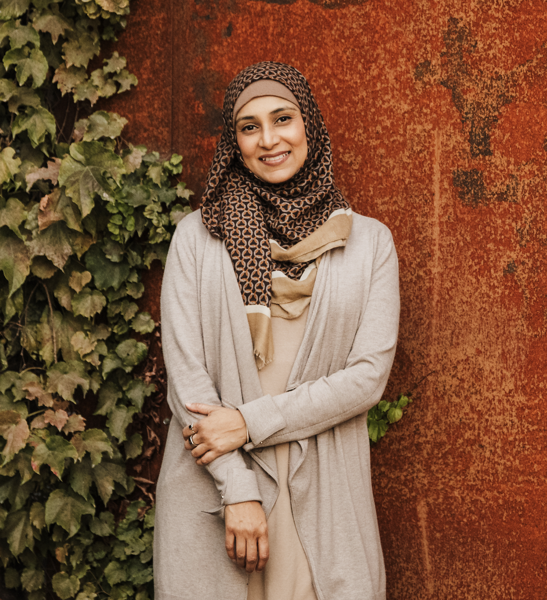 Smiling female doctor wearing a patterned hijab and beige coat standing in front of a rust-colored wall and green foliage.