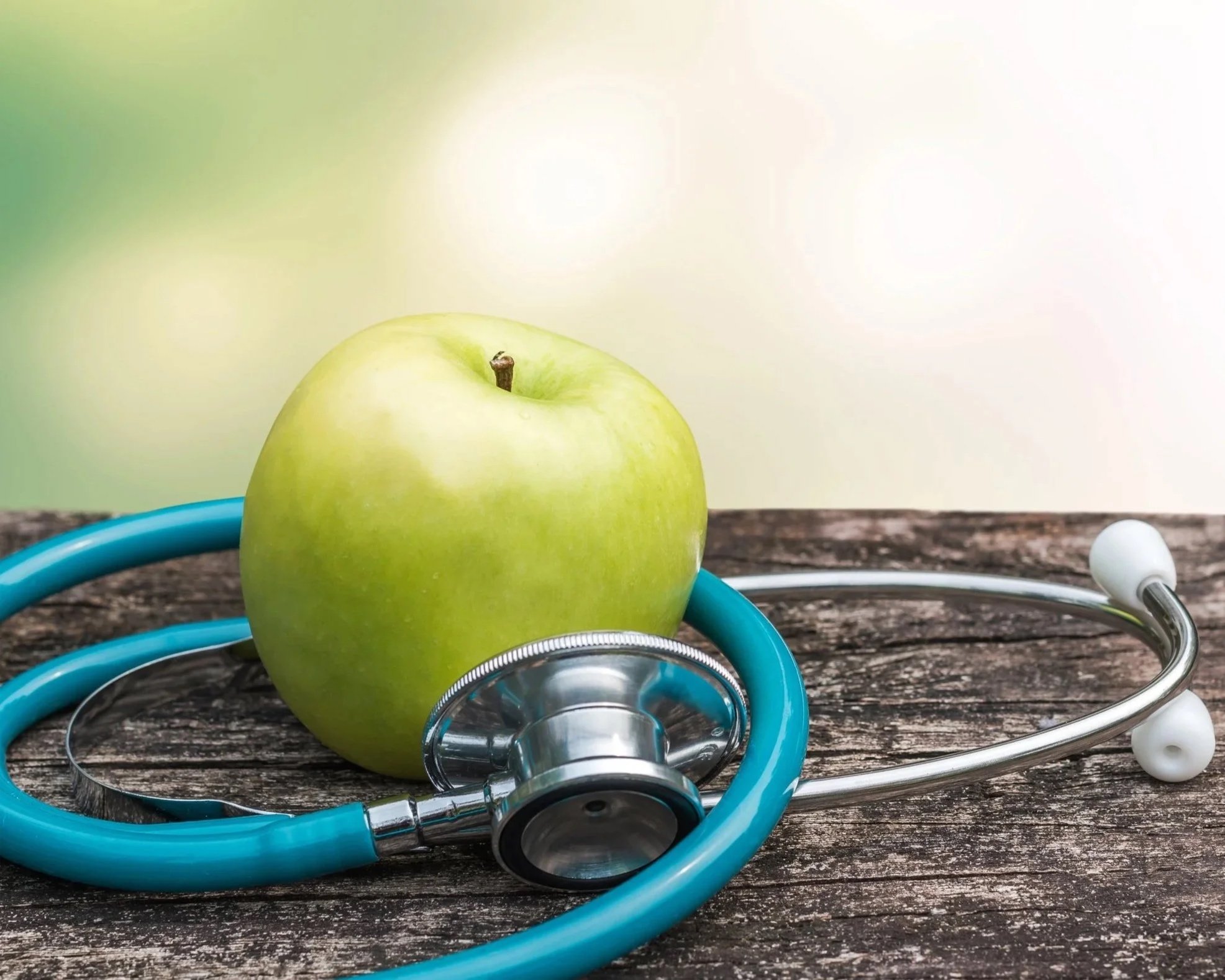 A green apple and a stethoscope on a wooden surface with a blurred natural background.