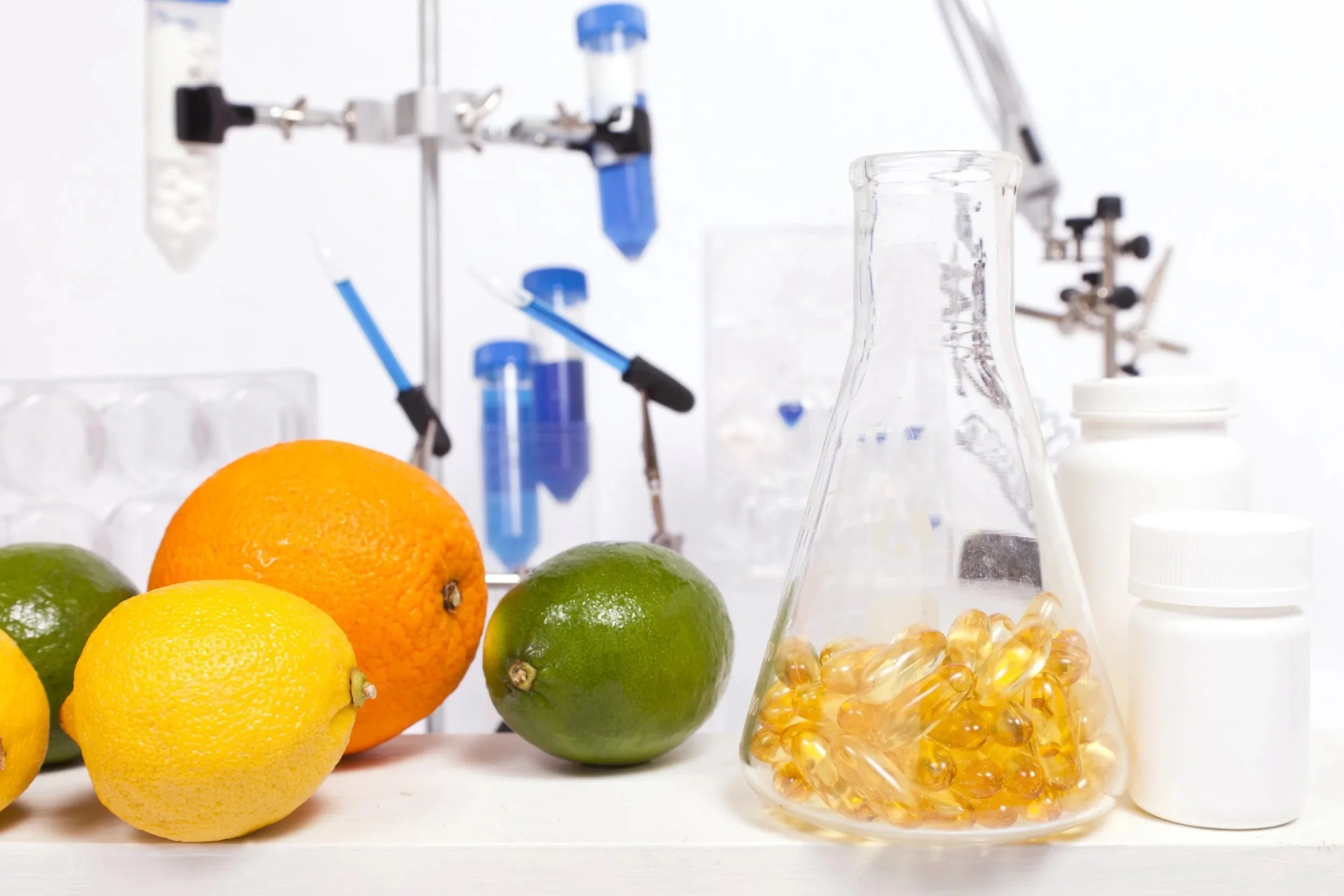 Various citrus fruits, including lemon, orange, and lime, along with a laboratory flask filled with yellow capsules or gel caps, and pharmaceutical bottles, set on a laboratory table with scientific equipment in the background.